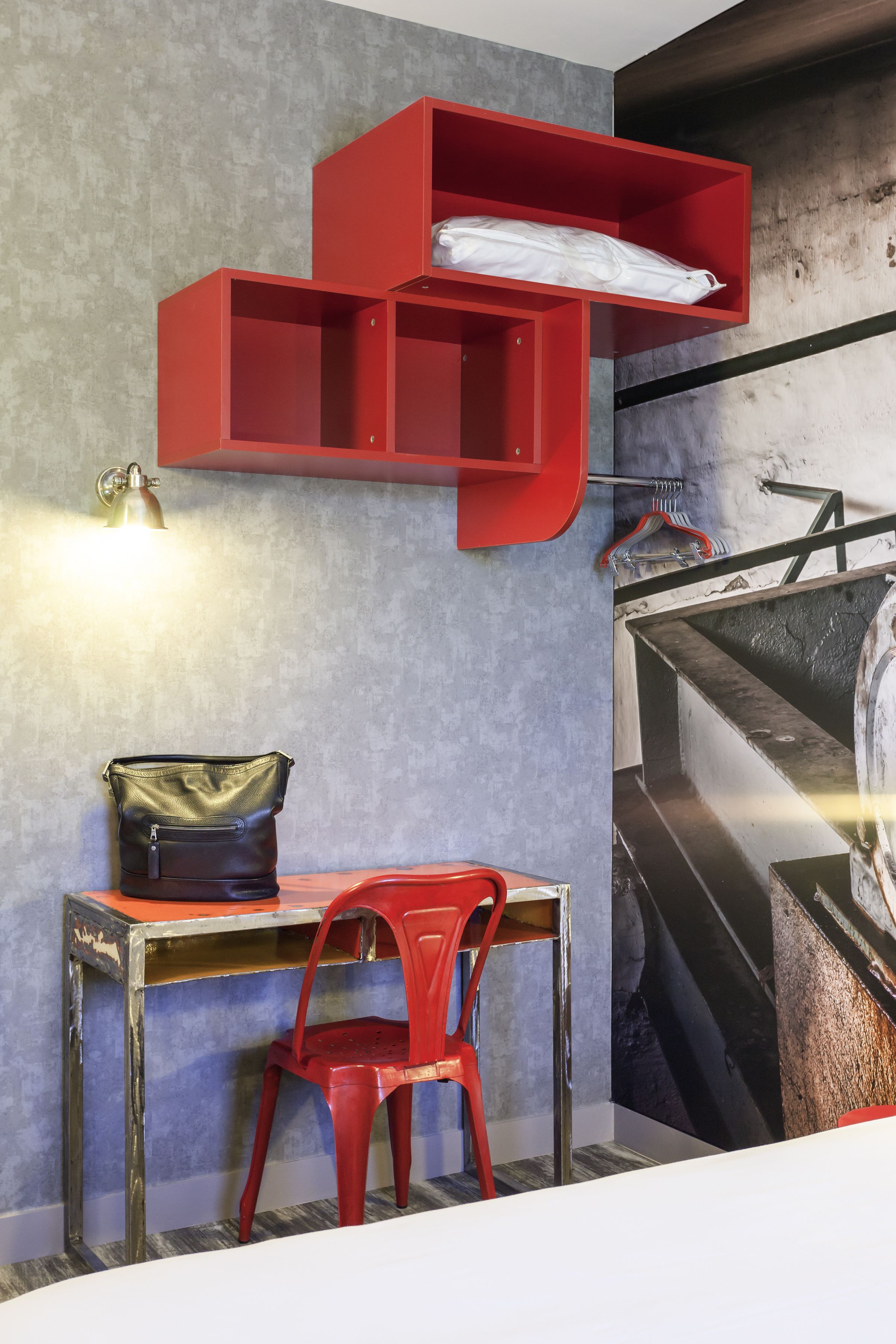 Una habitación con un escritorio, una silla roja, una estantería roja y una lámpara fijada a la pared.