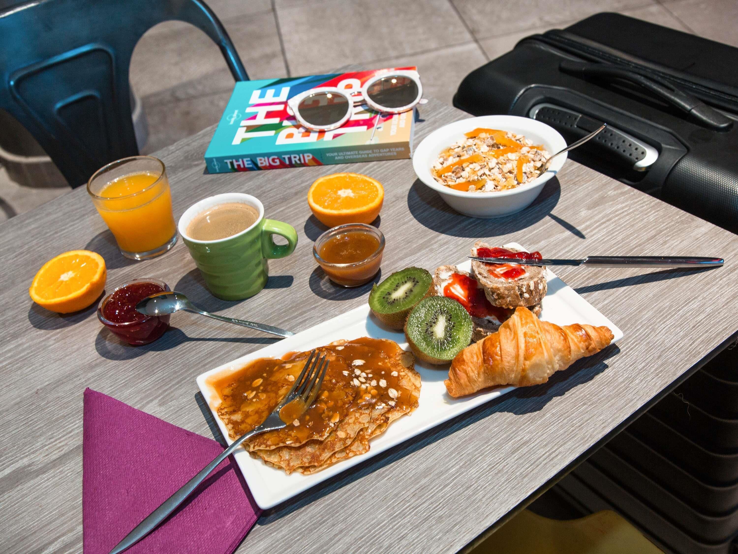 Ett frukostbord med en bok, en skål med flingor, en croissant och olika frukter.