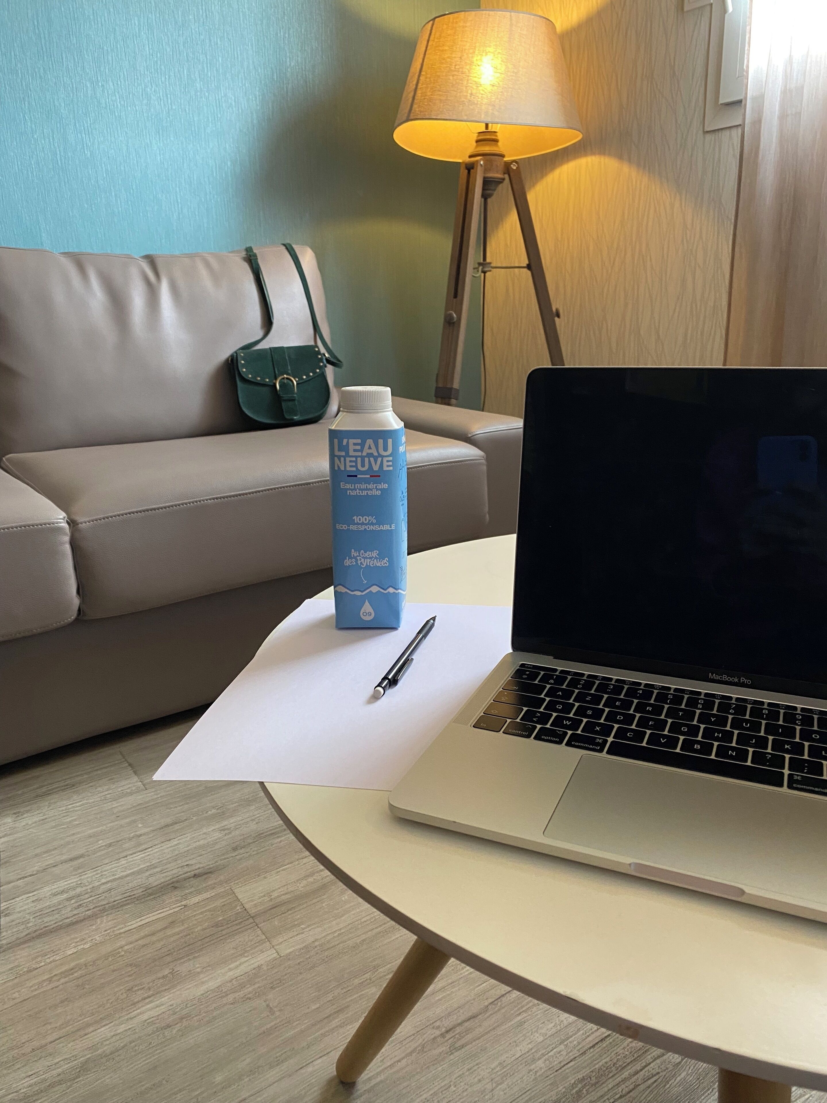 Una laptop sobre una mesa redonda, con una botella de agua y un bolígrafo. Un sofá gris y una lámpara se ven al fondo.