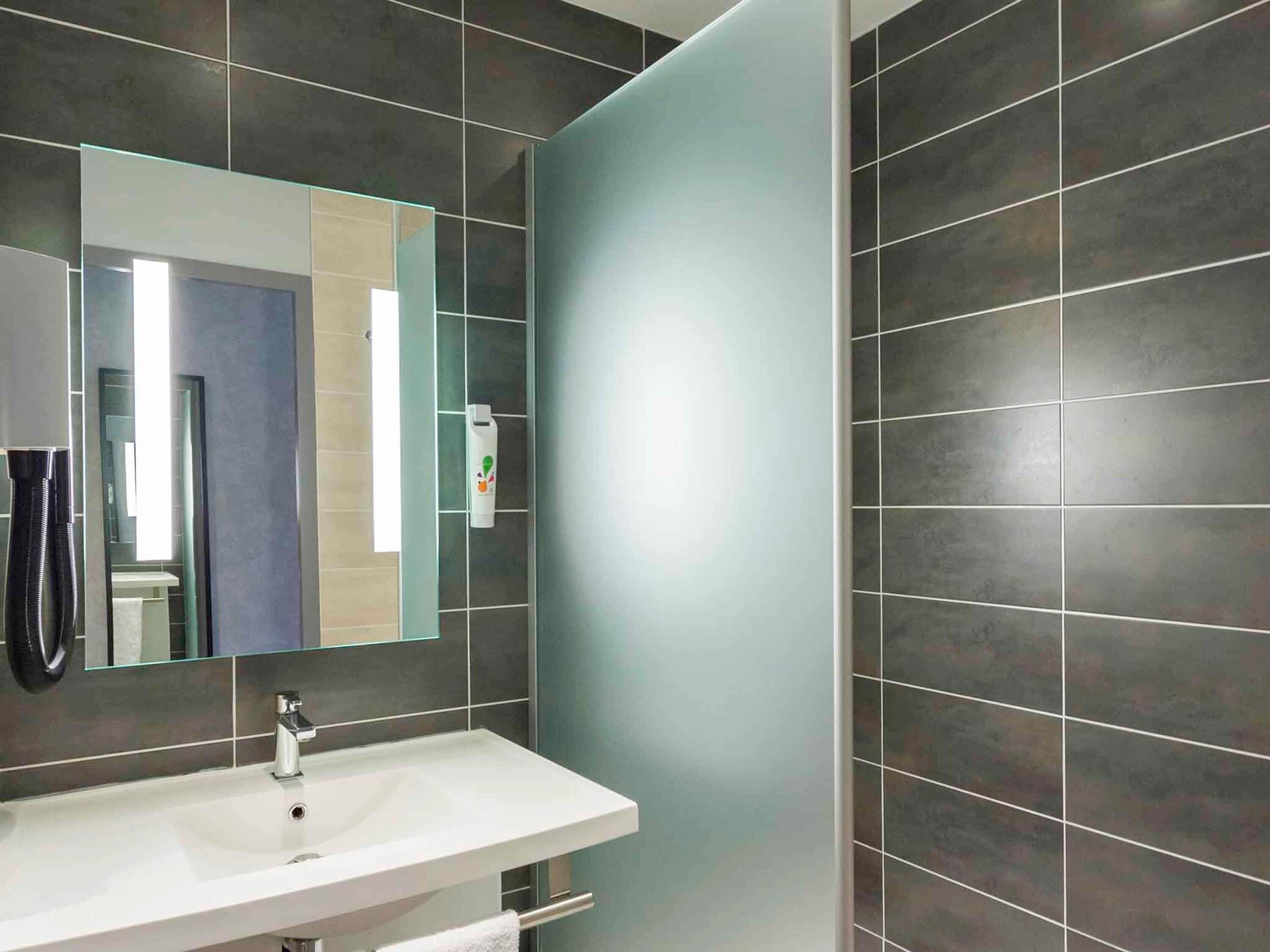 Une salle de bain moderne avec un lavabo blanc, un miroir et des murs recouverts de carreaux foncés.