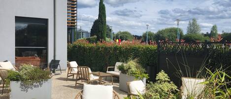 Een moderne buiten zithoek met een combinatie van rieten en gewatteerde stoelen, een ingebouwde plantenbak en uitzicht op een gebouw met grote ramen en een verticale houten panelenwand.