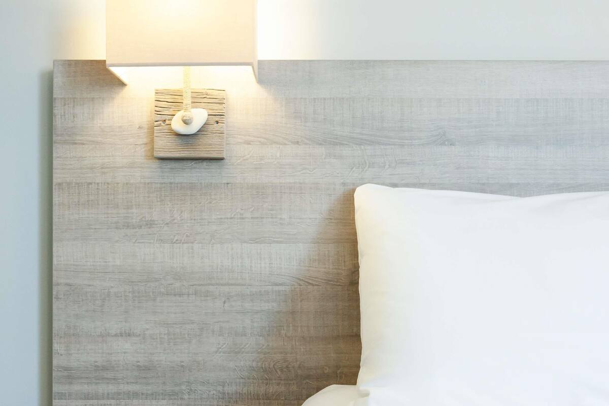 A bed with white bedding, a wooden headboard, and a wall-mounted light fixture.