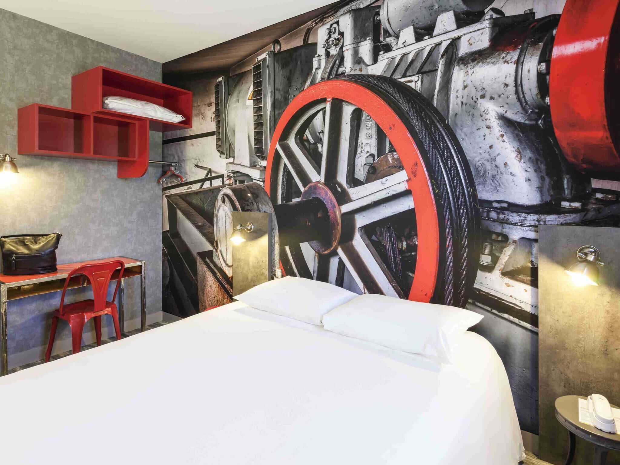 A hotel room with a large bed, a desk with a chair, a red shelf, and a wall mural of industrial machinery.
