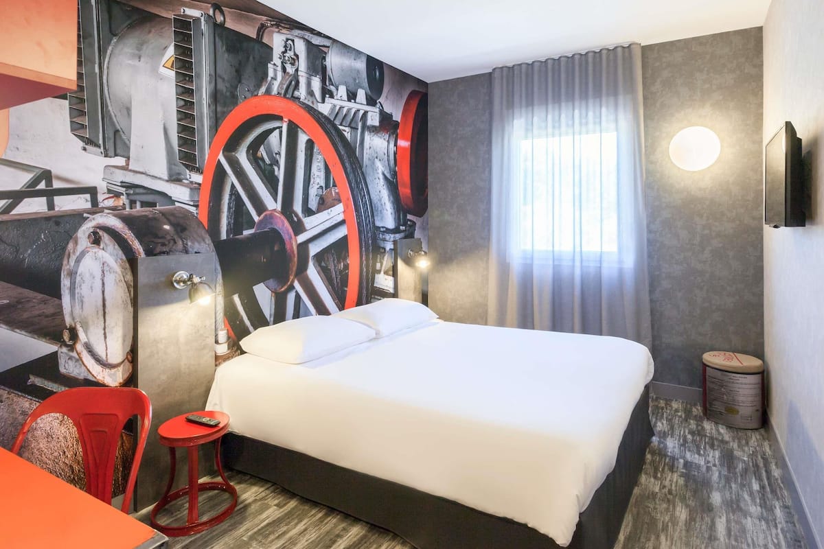 A hotel room with a bed, a red chair, a small table, a wall-mounted TV, and a large window with curtains.