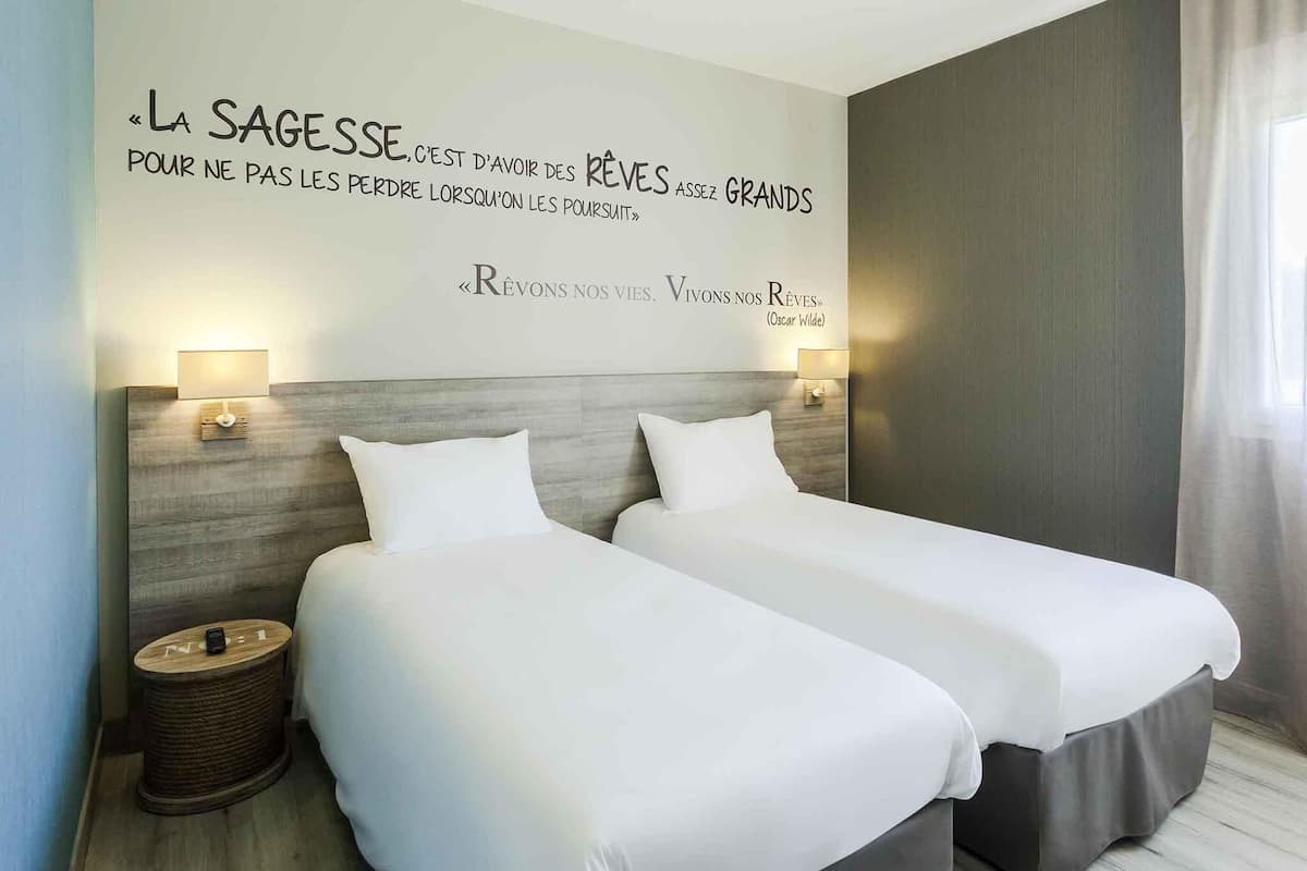 A hotel room with two beds, a quote on the wall, and a lamp on each side.