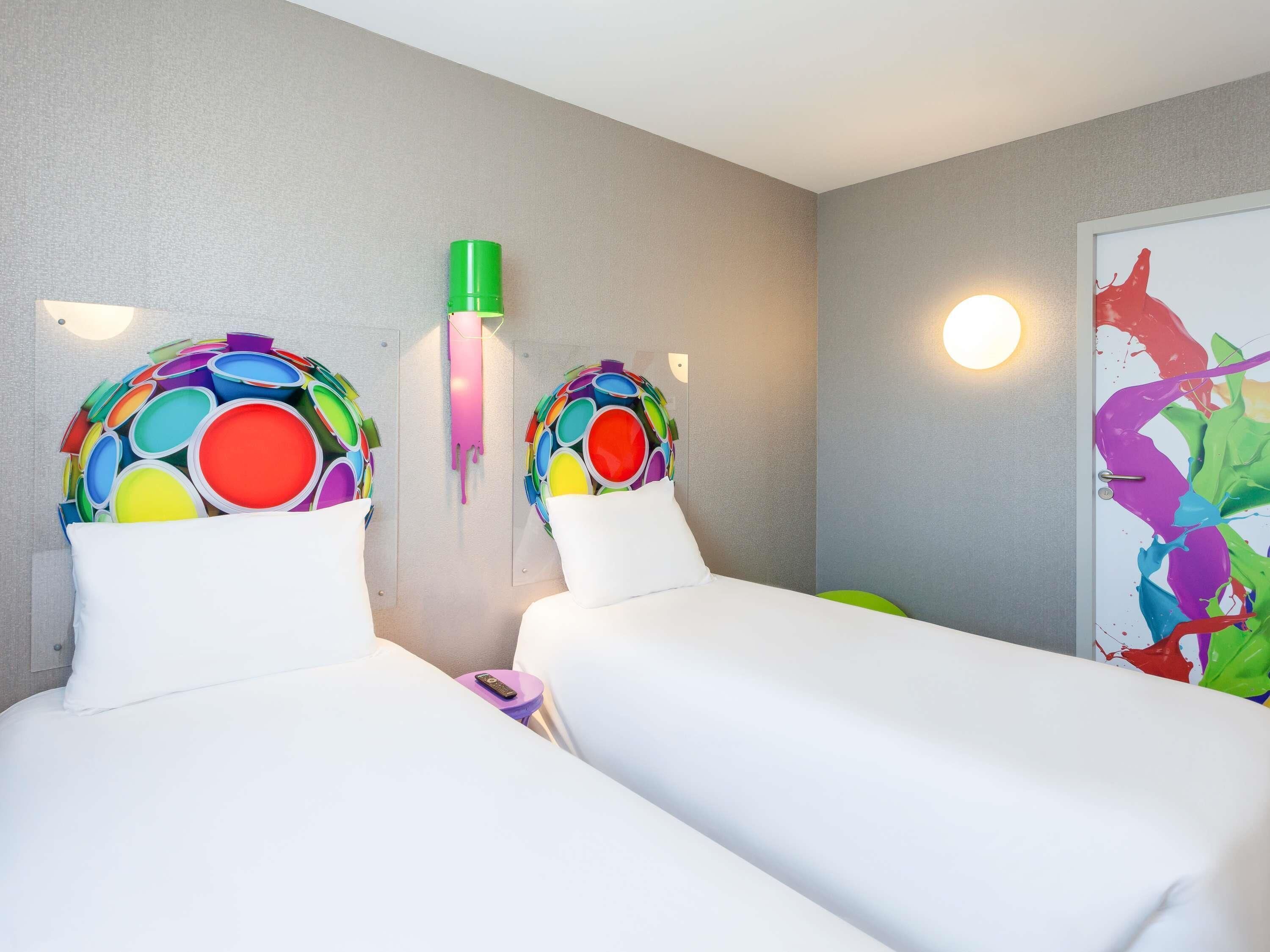 A hotel room with two beds, each with a colorful headboard, a wall-mounted light, and a painting on the wall.