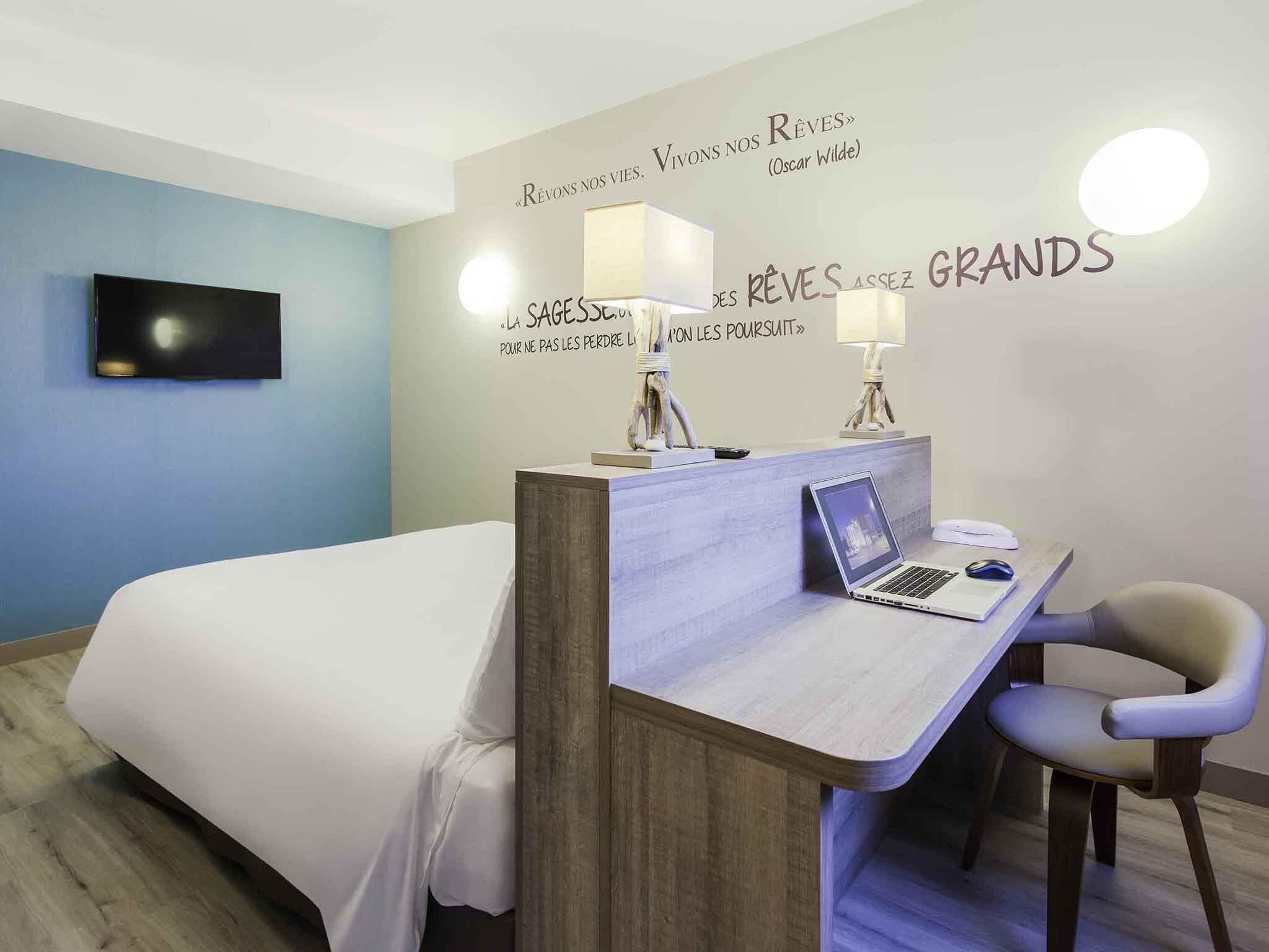 Habitación de hotel con cama, escritorio, computadora portátil, una silla y decoración en la pared con texto en francés.