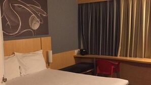 A hotel room with a bed, a desk, a chair, a window with curtains, and a wall art piece.