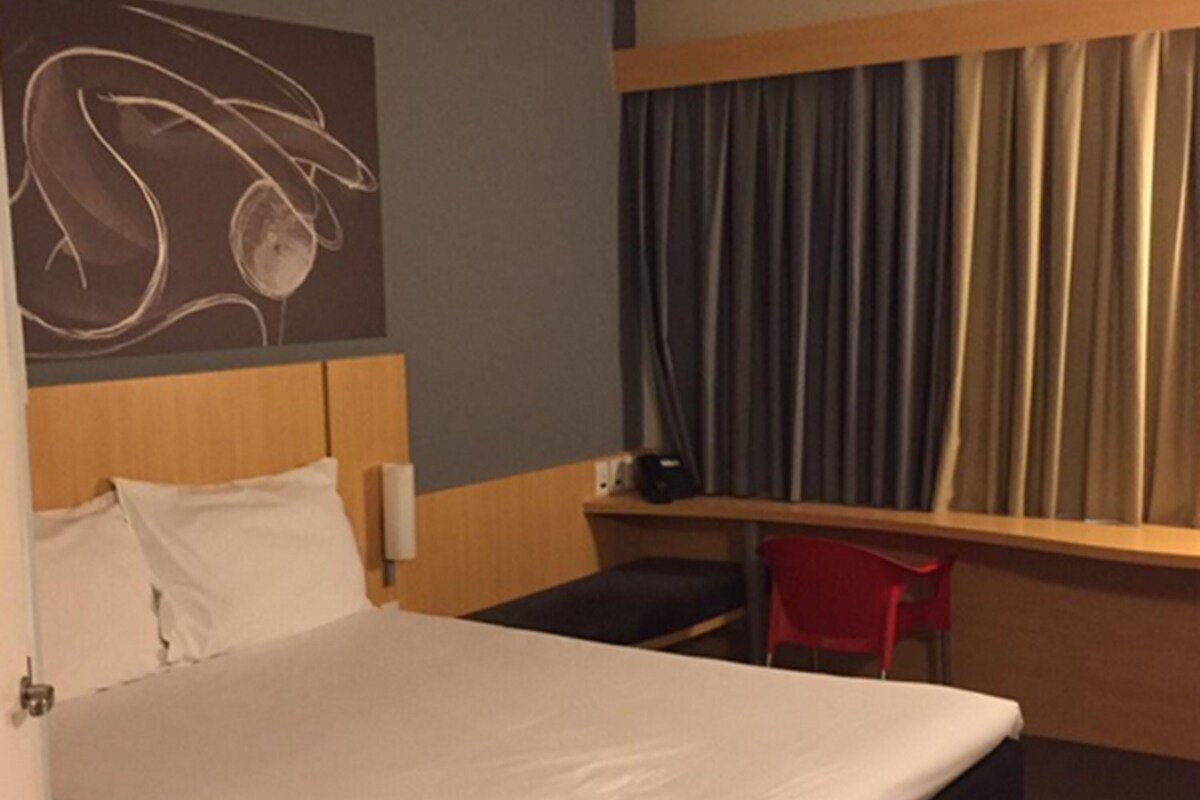 A hotel room with a bed, a desk, a chair, a window with curtains, and a wall art piece.