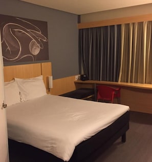 A hotel room with a bed, a desk, a chair, a window with curtains, and a wall art piece.