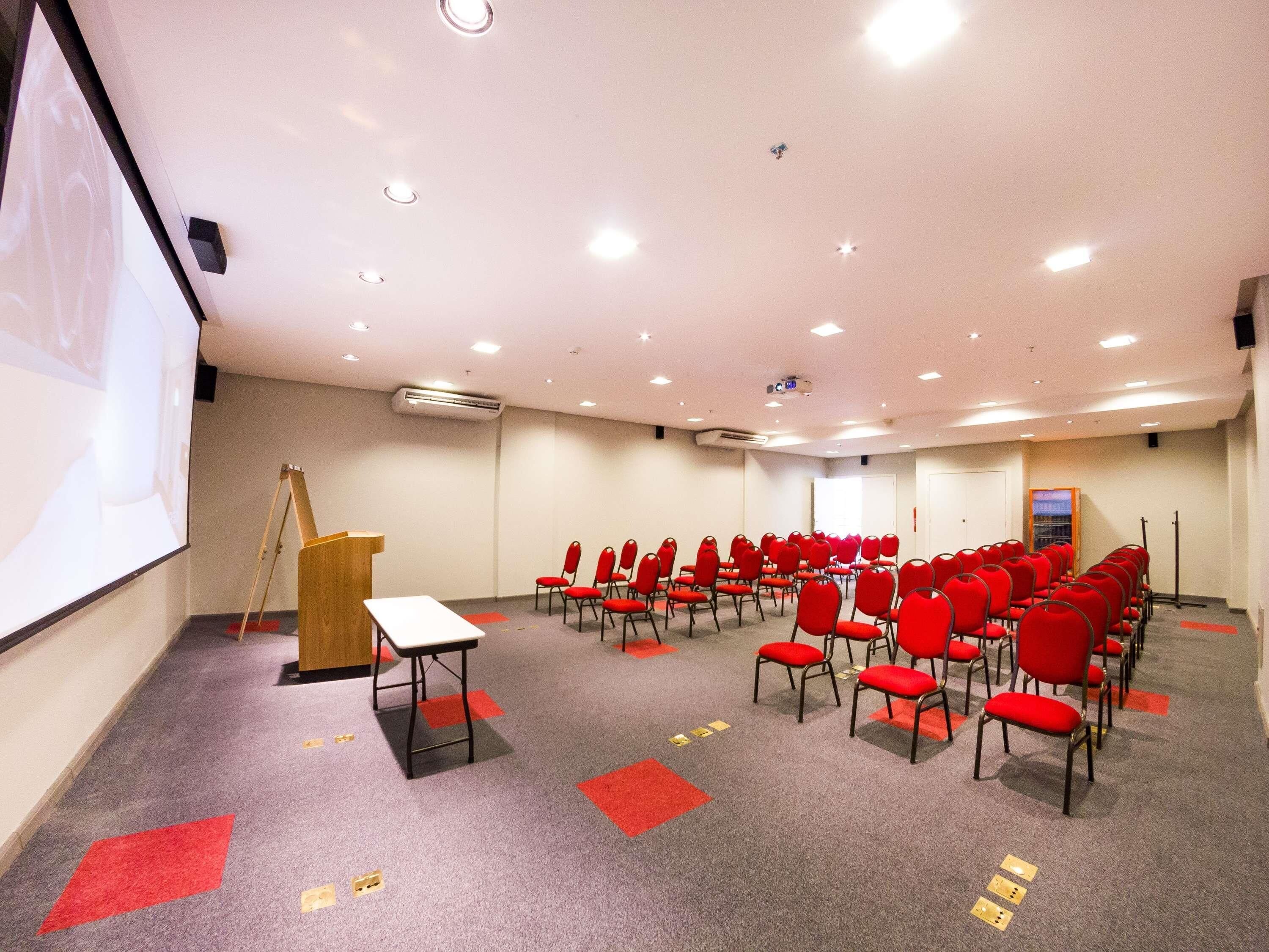 A conference room with rows of red chairs, a podium, and a projector screen.
