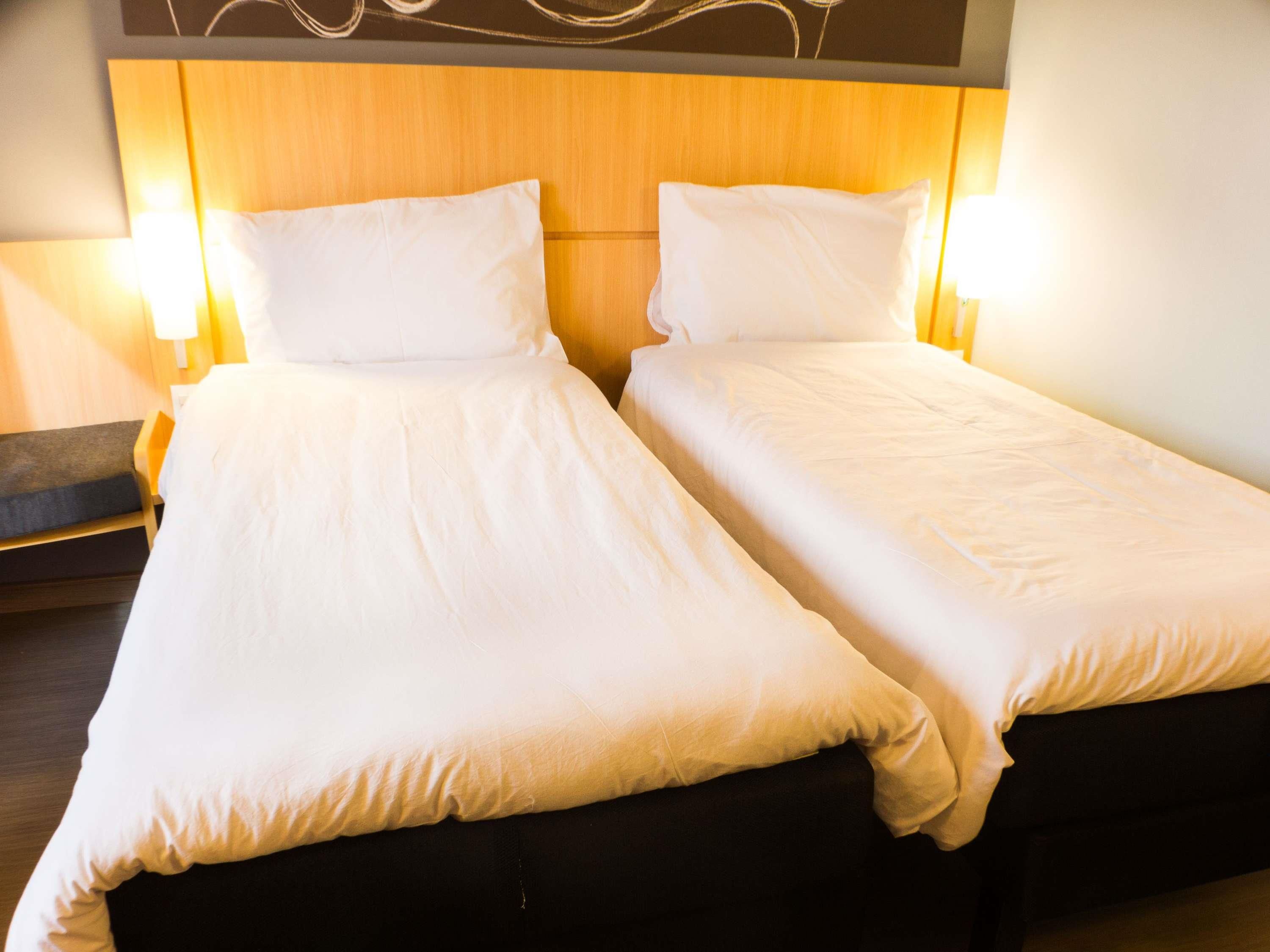 Un lit double avec du linge de lit blanc et une tête de lit en bois, flanqué de deux lampes fixées au mur.