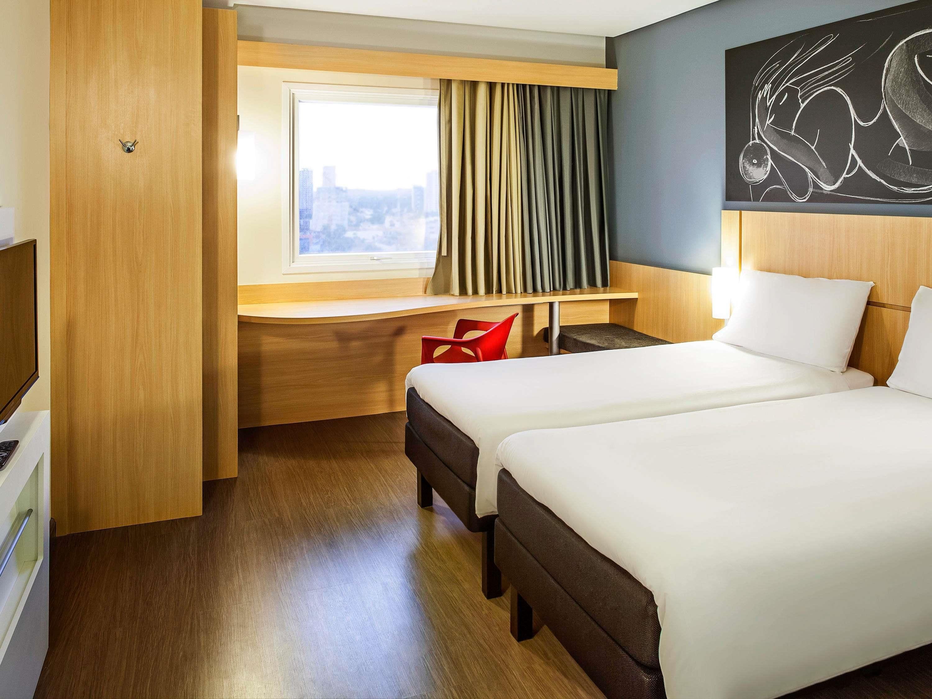 A hotel room with two beds, a desk, a red chair, and a city view.