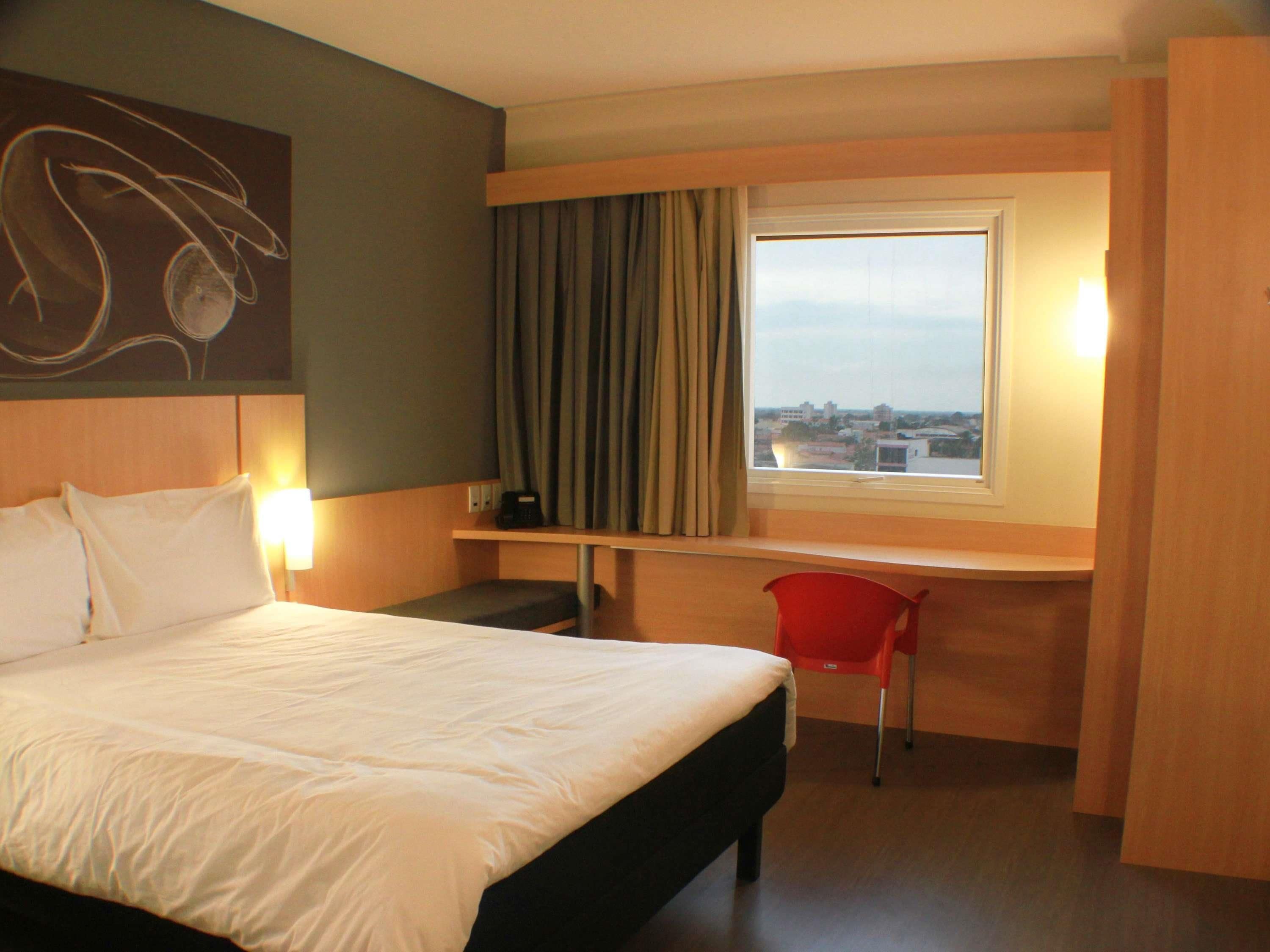A hotel room with a bed, a desk, a chair, a window with a view, and a wall with a light fixture.