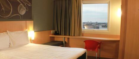 A hotel room with a bed, a desk, a chair, a window with a view, and a wall with a light fixture.