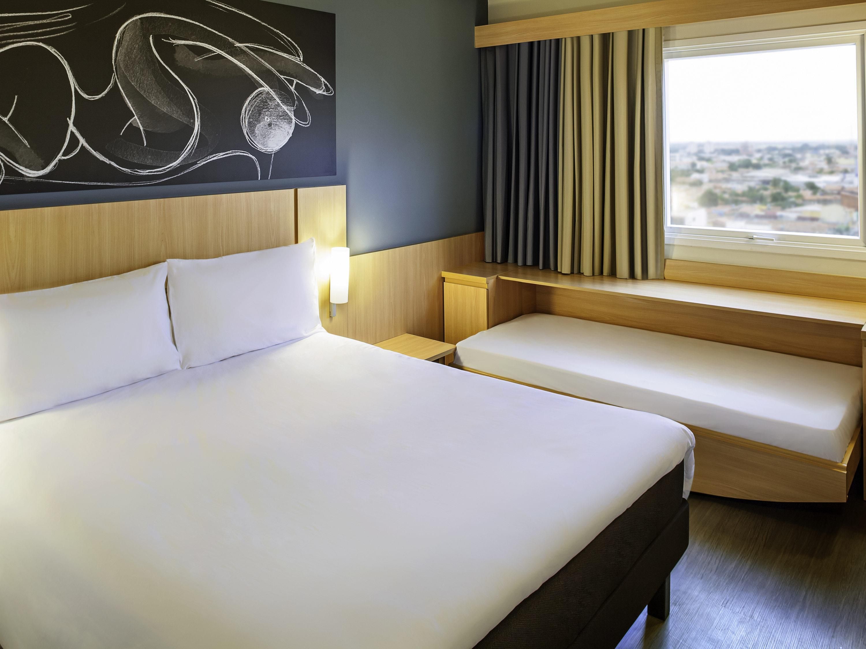 Une chambre d’hôtel avec un grand lit, une table de chevet, un petit bureau, une fenêtre avec des rideaux et une œuvre d’art accrochée au mur.