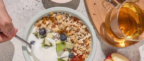 A bowl of yogurt with fruits and granola, a glass of amber liquid, and sliced bread on a table.