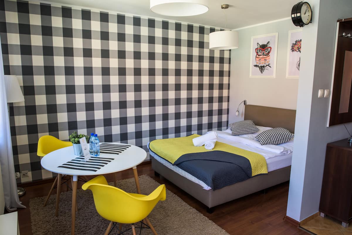 A hotel room with a checkered wall, a bed with a yellow blanket, a bedside table with a lamp, and a small plant.