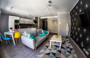 Una sala moderna con un sofá gris, sillas coloridas, una mesa de centro blanca y una cocina al fondo.