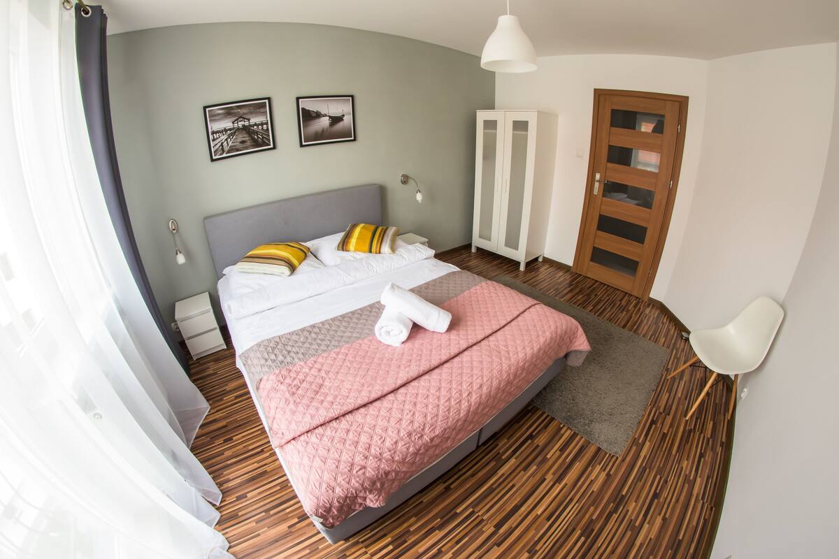A neatly made bed with a pink quilt, a wooden floor, a white wardrobe, and two framed pictures on the wall.