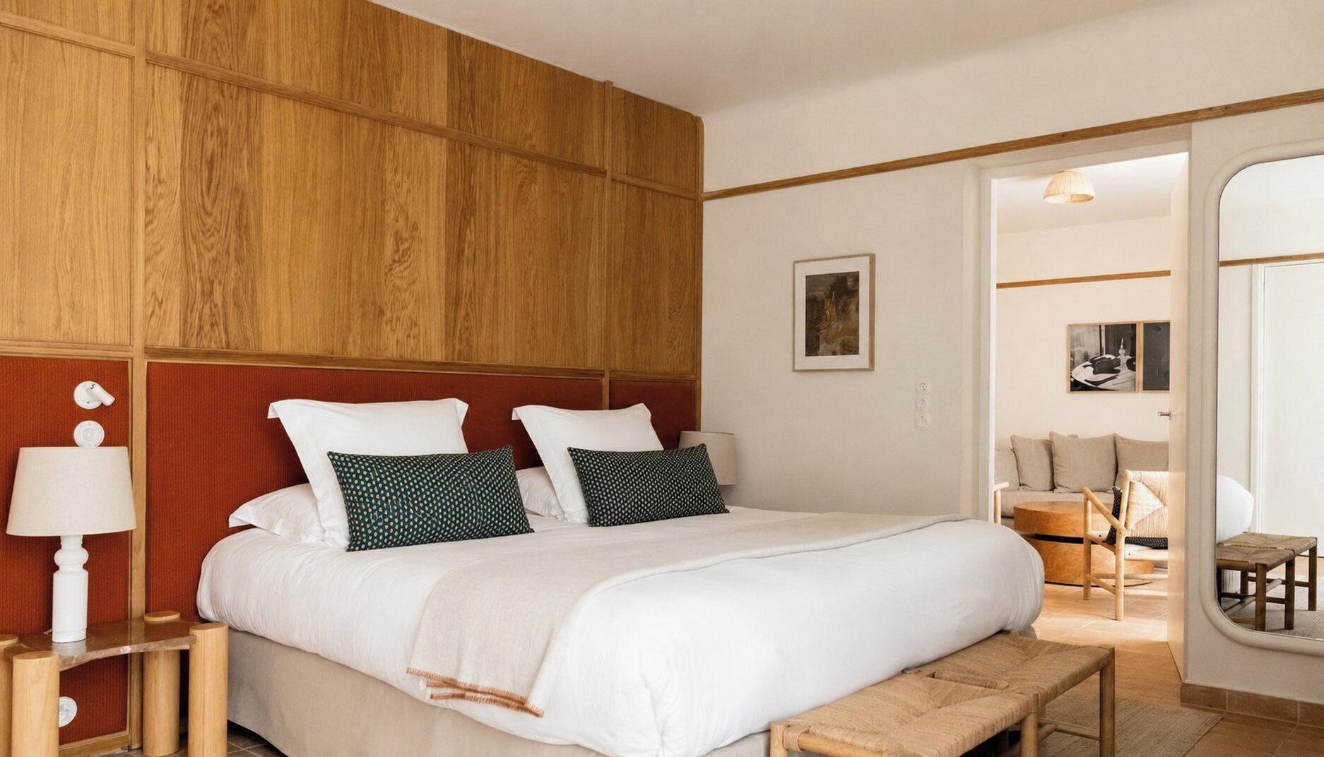 Habitación de hotel con cabecera de madera, cama con ropa blanca, mesita de noche con lámpara y vistas al área de estar contigua.
