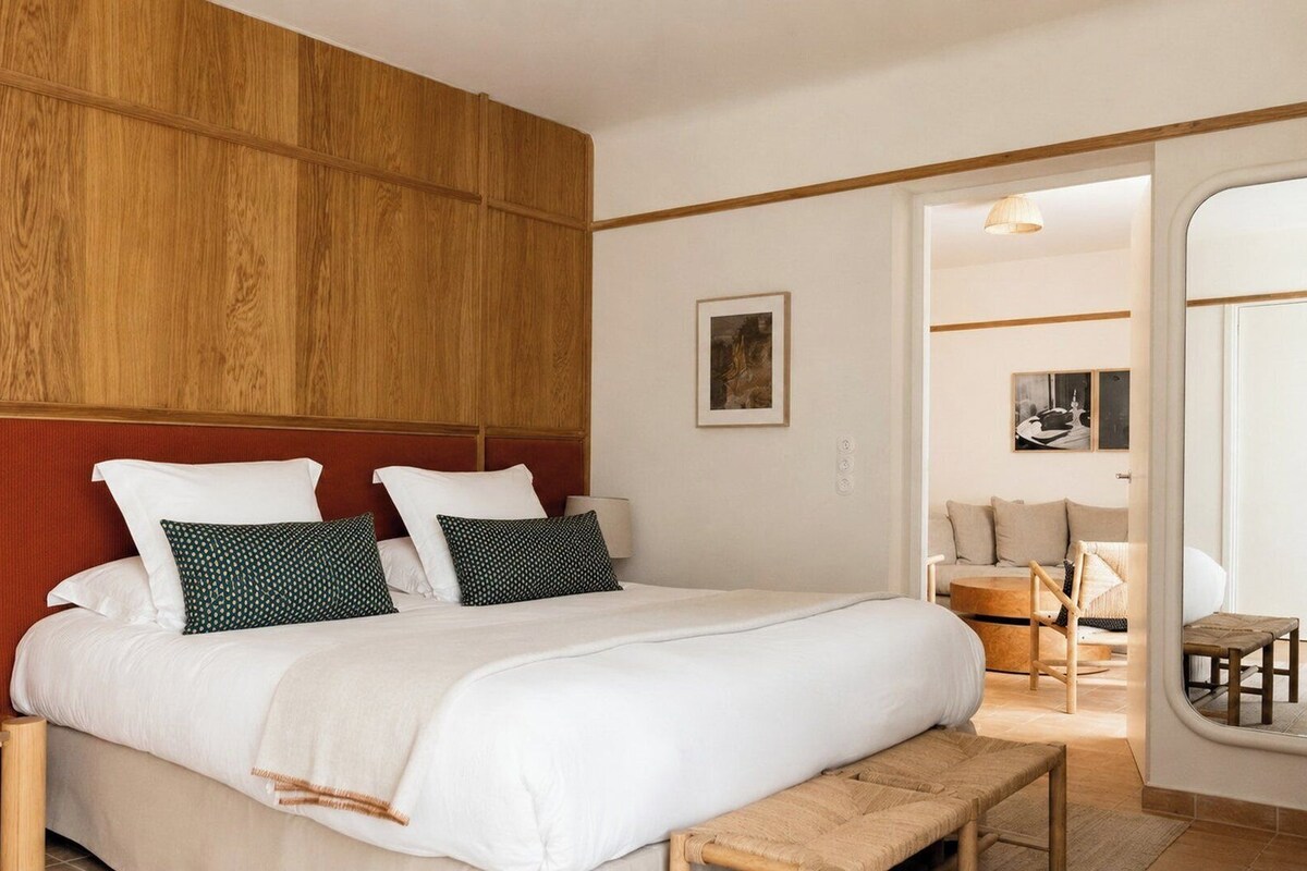 A hotel room with a wooden headboard, a bed with white bedding, a bedside table with a lamp, and a view into the adjacent living area.