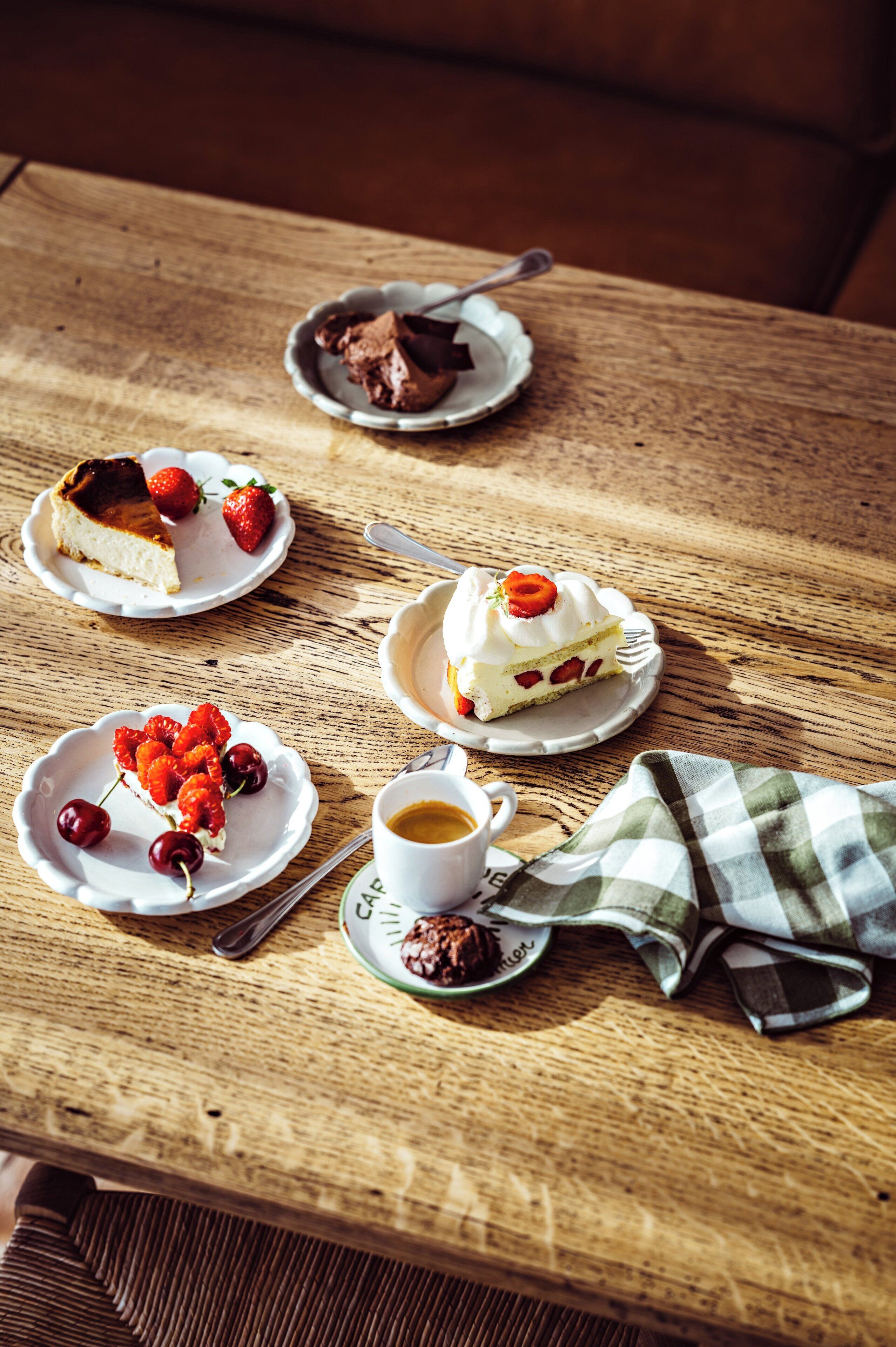 Une table en bois sur laquelle sont disposées des assiettes de desserts, notamment une part de gâteau au fromage, un gâteau aux fraises et un dessert à la crème fouettée et aux fraises. On y trouve également une tasse de café et une serviette en tissu à carreaux verts.