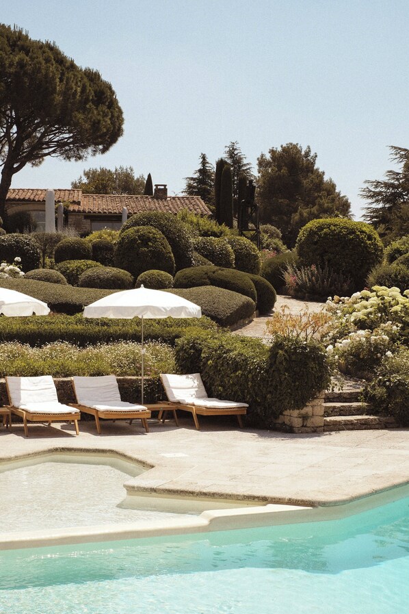 Un área de piscina con sillones de descanso y sombrillas, rodeada de jardines cuidados y un árbol grande.