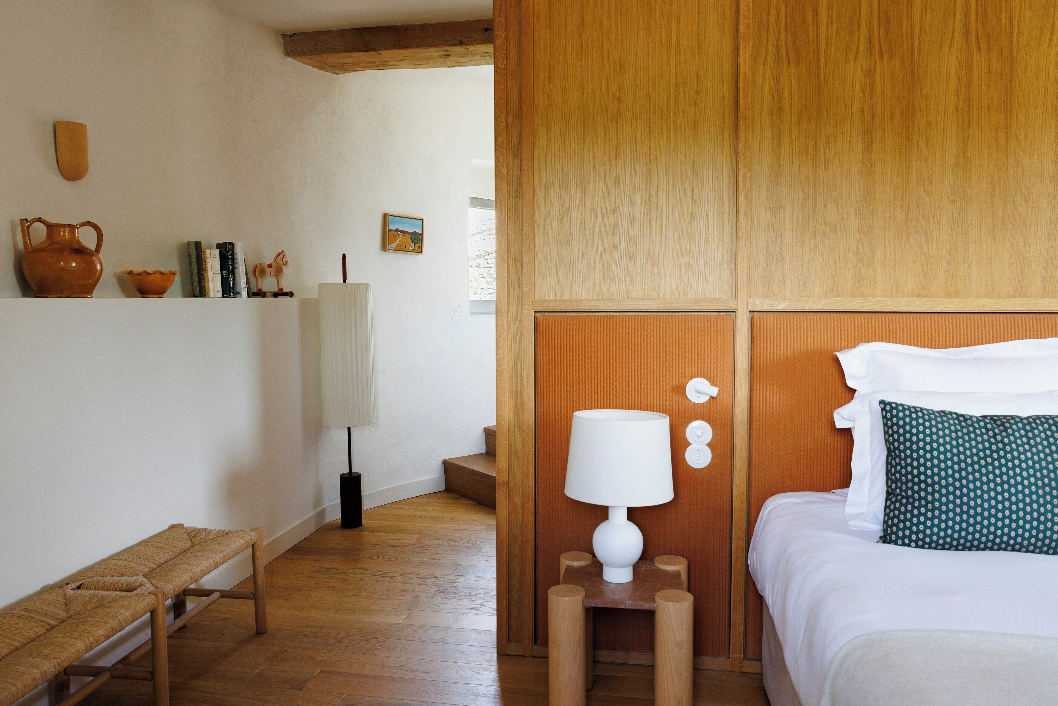 A room with a bed, a bench, a lamp, and a shelf with books and decorative items.