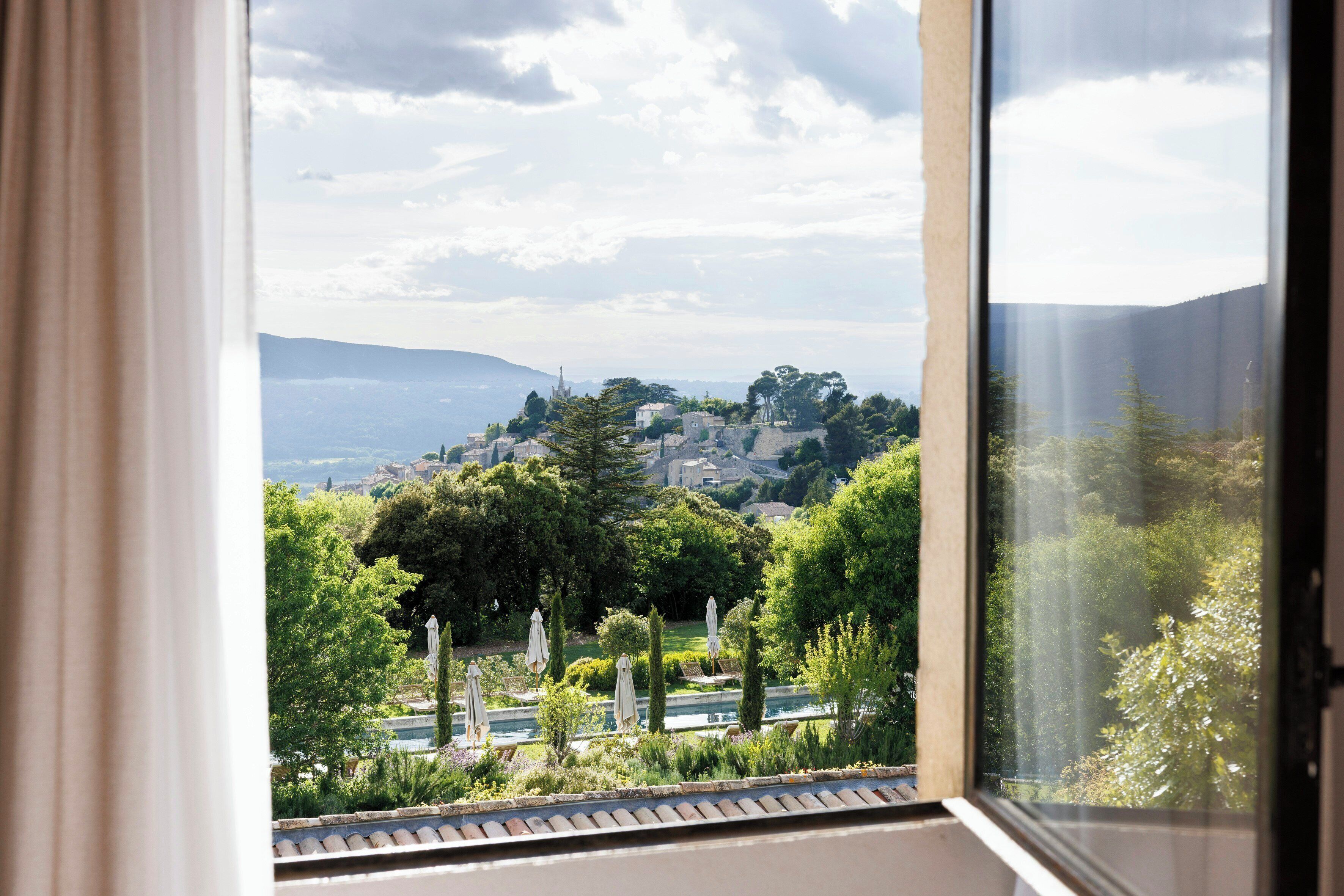 Vista por una ventana que muestra un paisaje con árboles, una piscina y una colina distante.