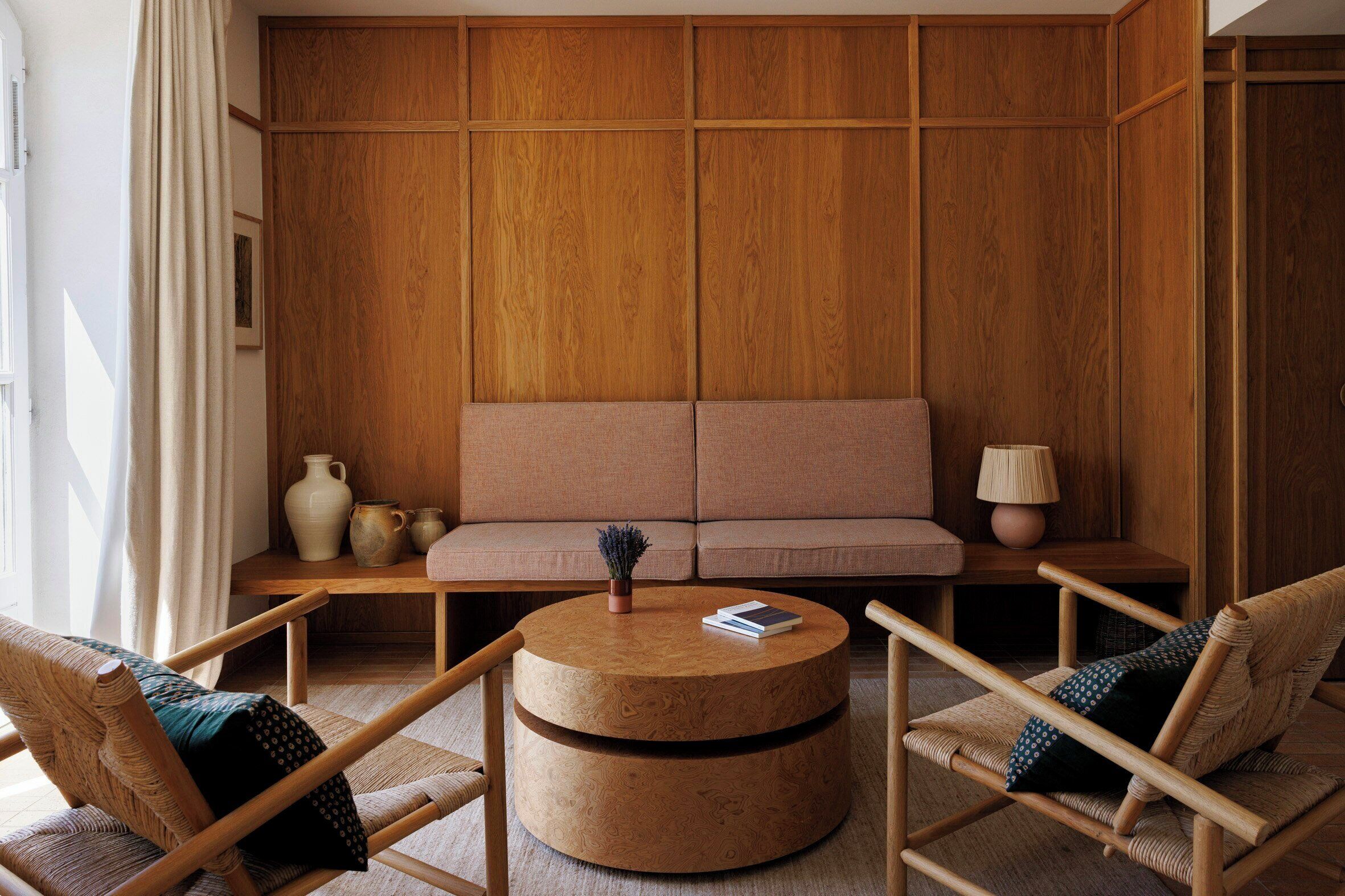 A room with a wooden panel wall, a sofa, two armchairs, a round wooden coffee table, and a small side table with a lamp.