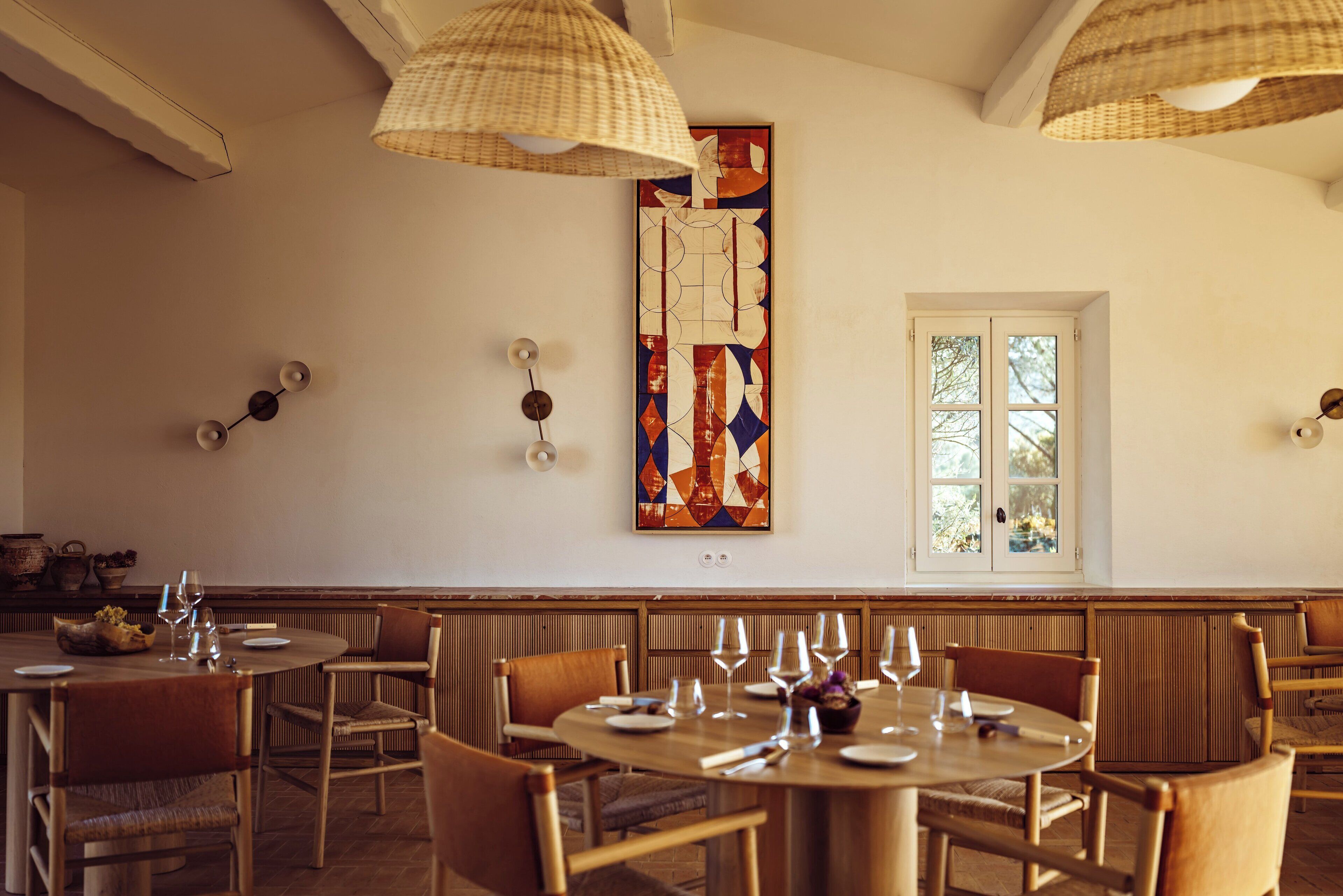 A dining area with round tables and wooden chairs, a decorative wall piece, and a window with a view.