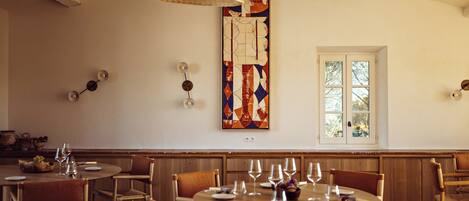 A dining area with round tables and wooden chairs, a decorative wall piece, and a window with a view.
