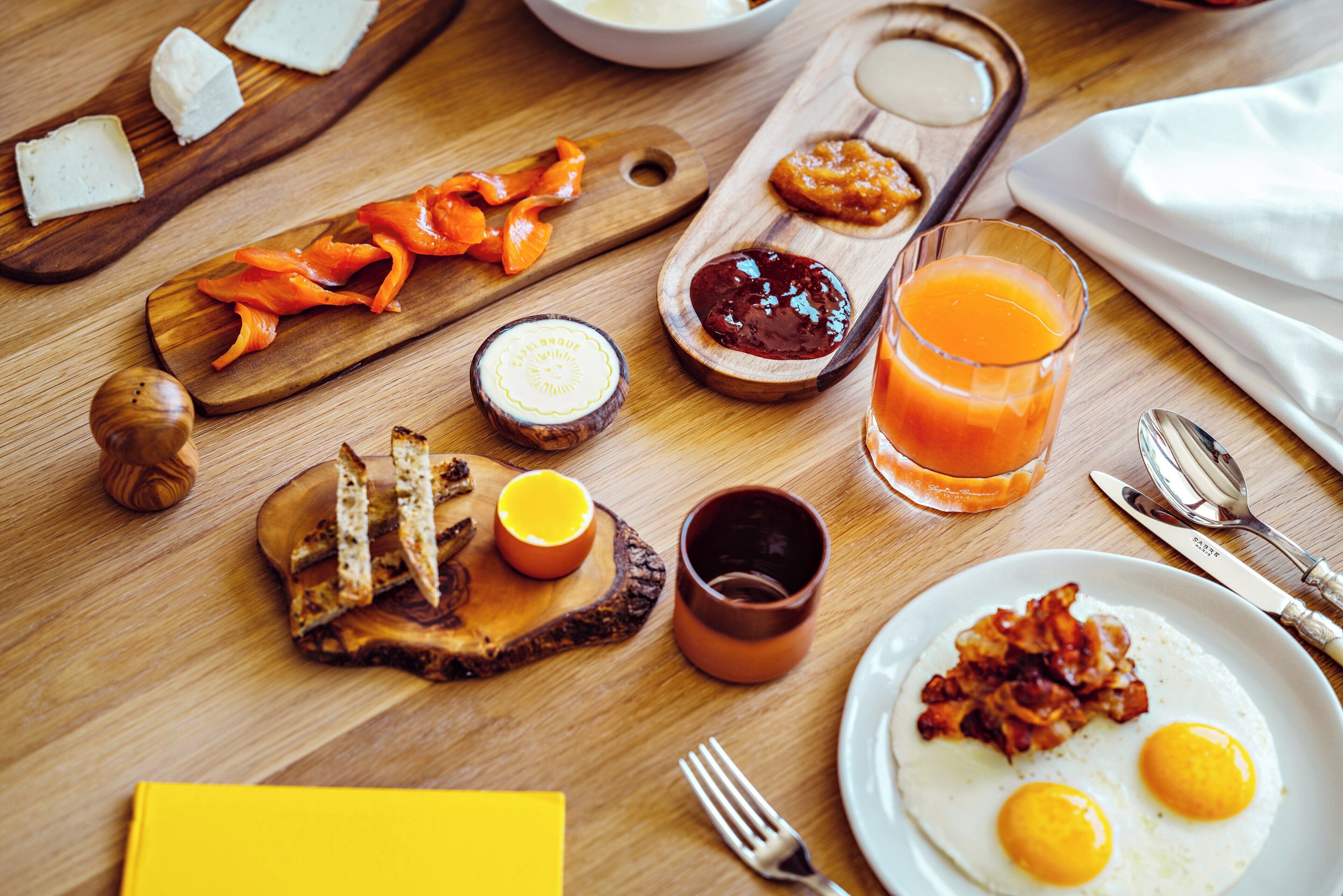 Desayuno con tostadas, huevos, tocino, jugo de naranja y diferentes condimentos sobre una mesa de madera.