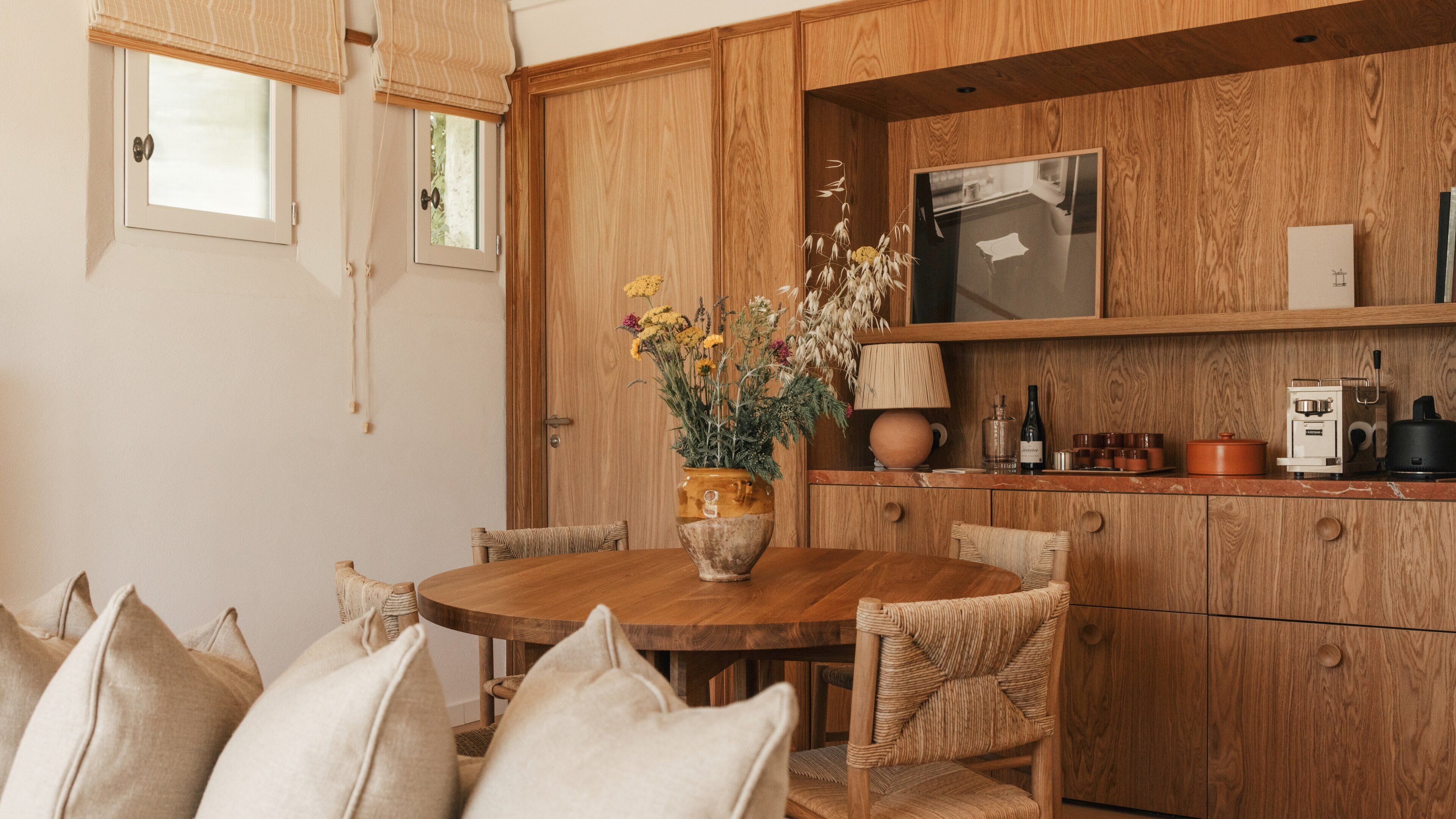 A wooden dining table with chairs, a vase with flowers, a kitchen area with wooden cabinets, and a window with blinds.