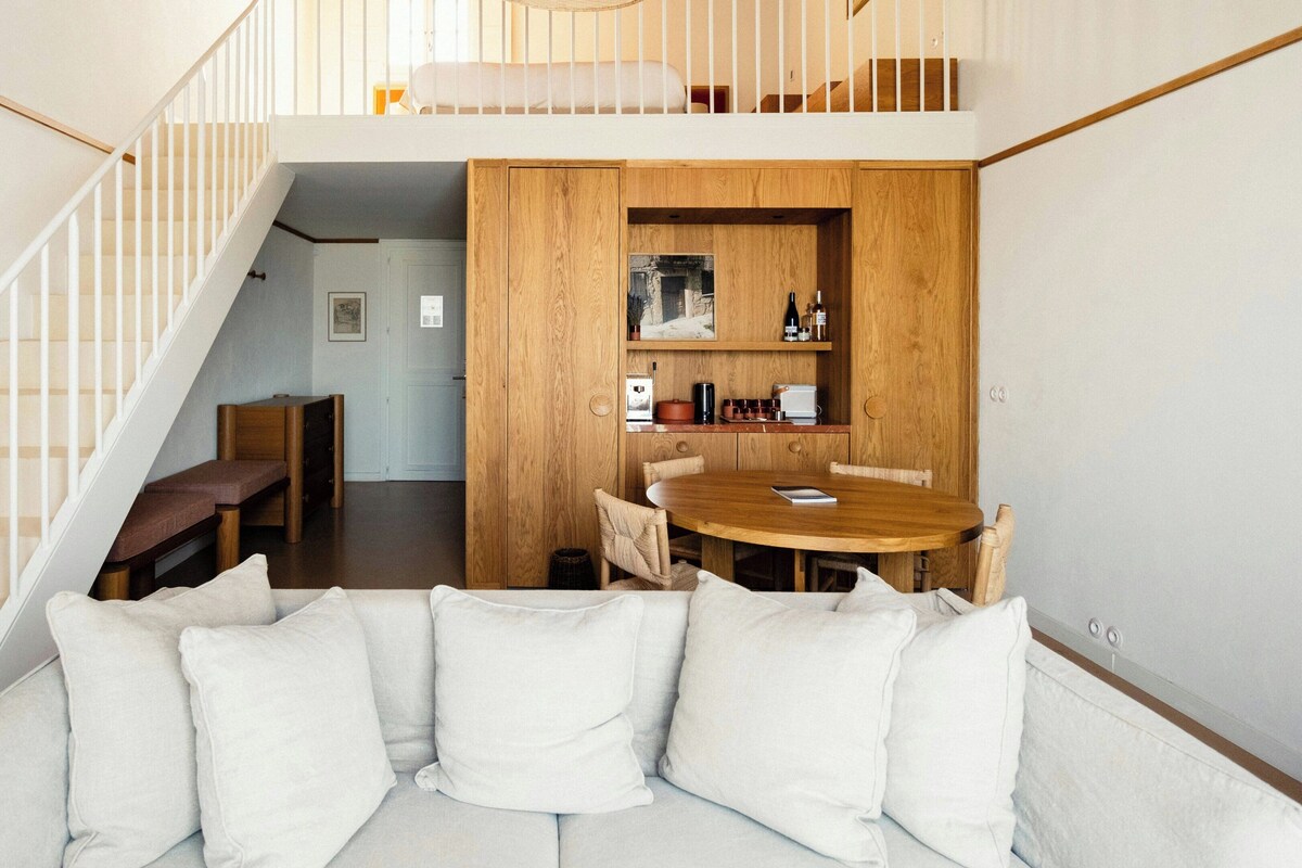 A modern loft-style living space with a staircase, a wooden dining table, and a sofa with cushions.