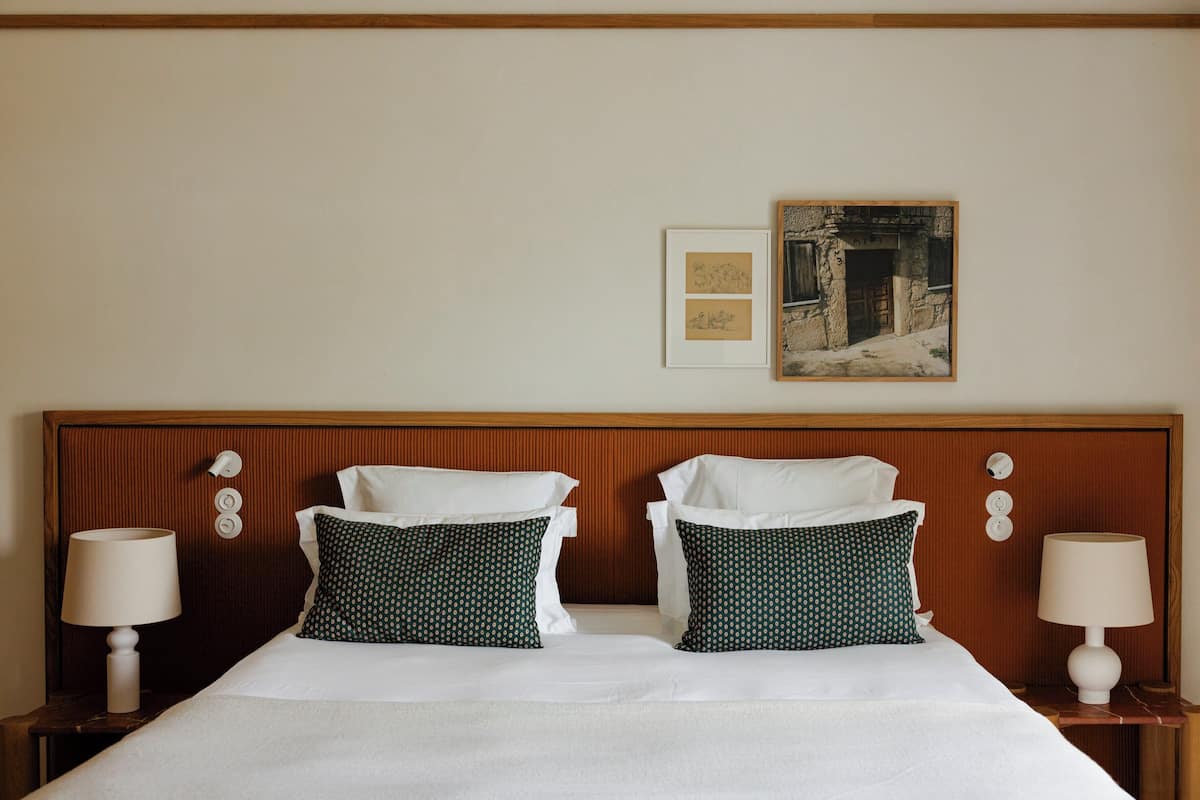 Habitación de hotel con cabecera de madera, dos almohadas, una cama, dos lámparas de noche y dos cuadros en la pared.