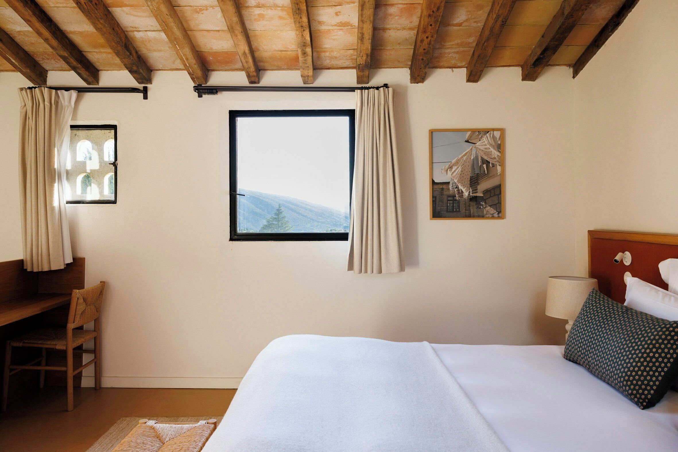 A bedroom with a bed, a desk, a chair, a window with curtains, a lamp, and a framed picture on the wall.