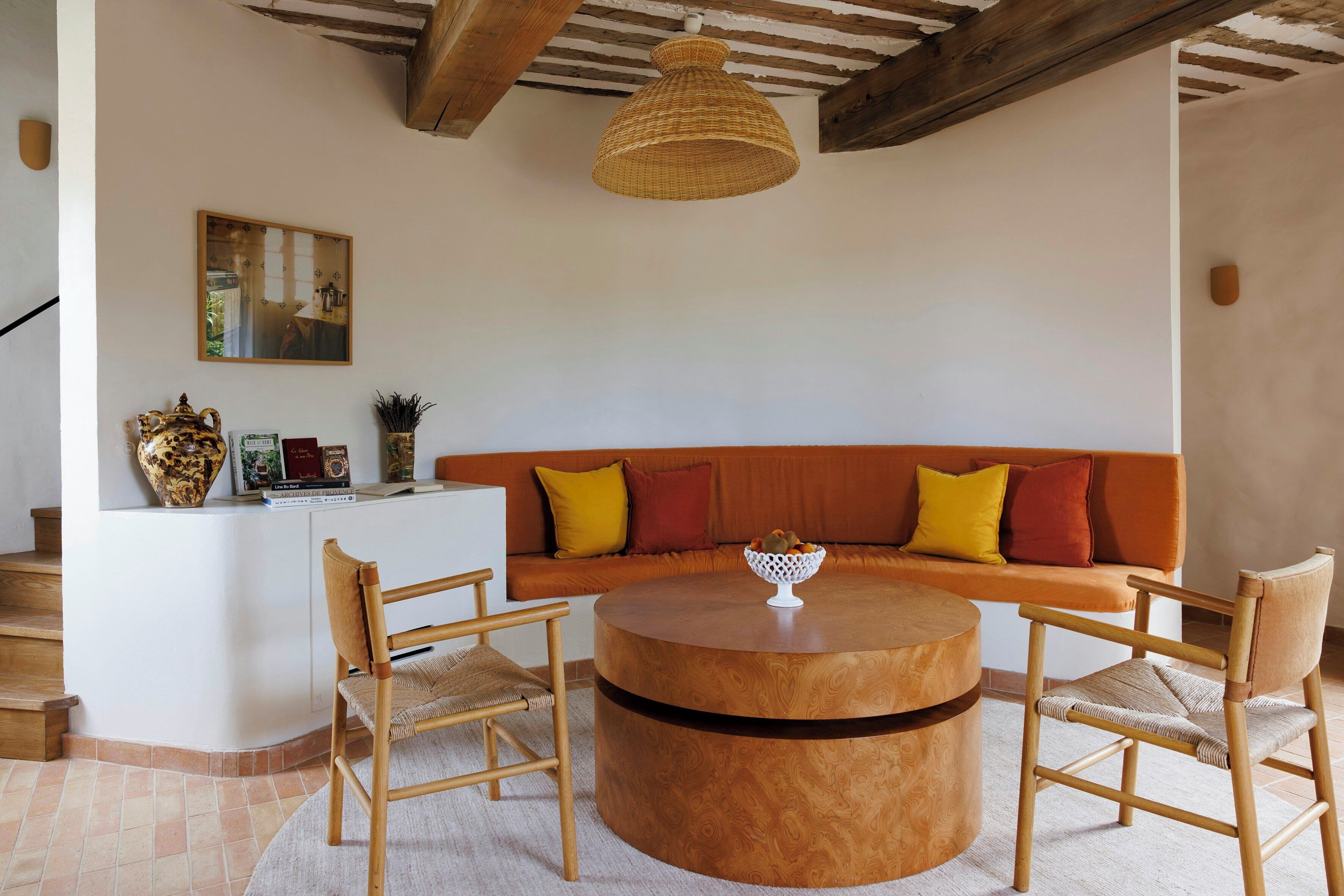 Une pièce avec une table ronde en bois, deux chaises, un canapé avec des coussins, un luminaire suspendu en fibres tressées et un tableau encadré au mur.