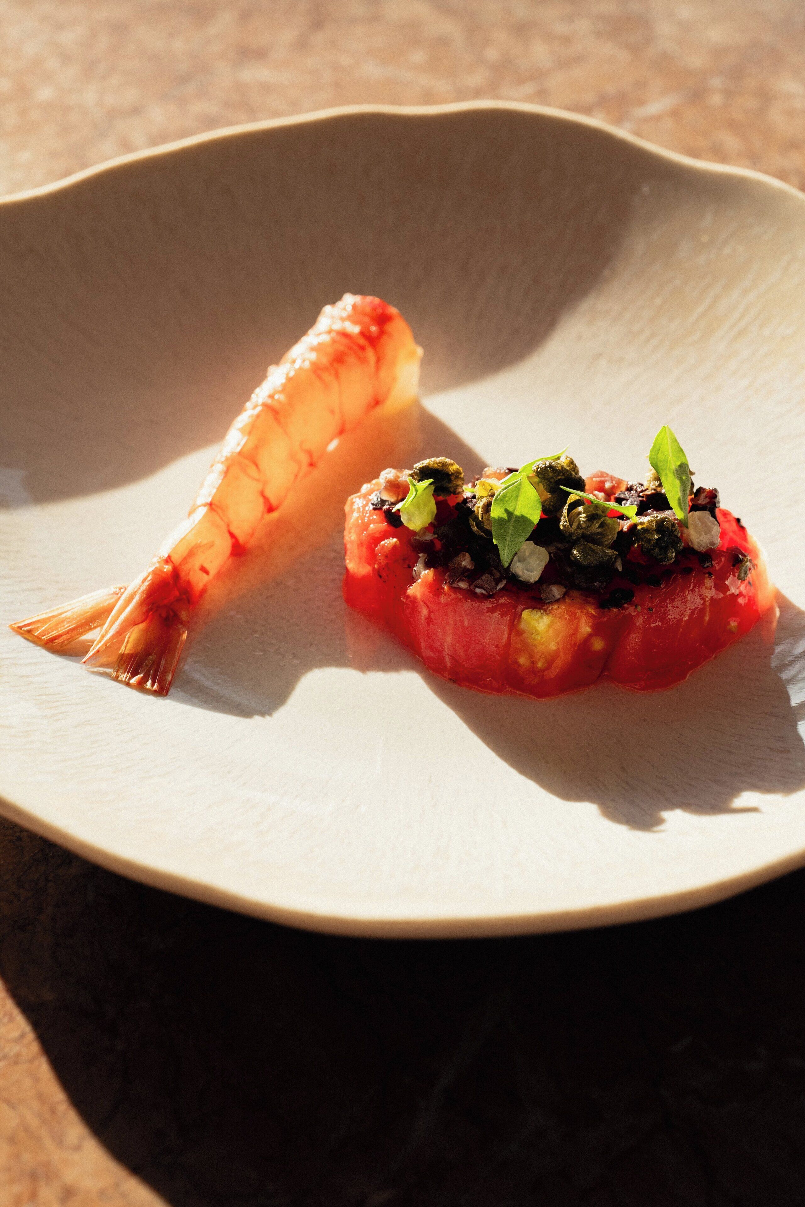 A plate with a shrimp and a tomato-based dish, possibly a tartare, on a textured surface.