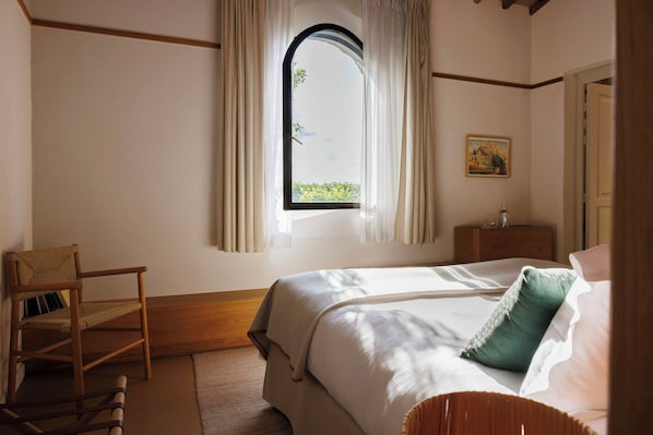 A bedroom with a bed, a chair, a window with curtains, a nightstand, and a painting on the wall.