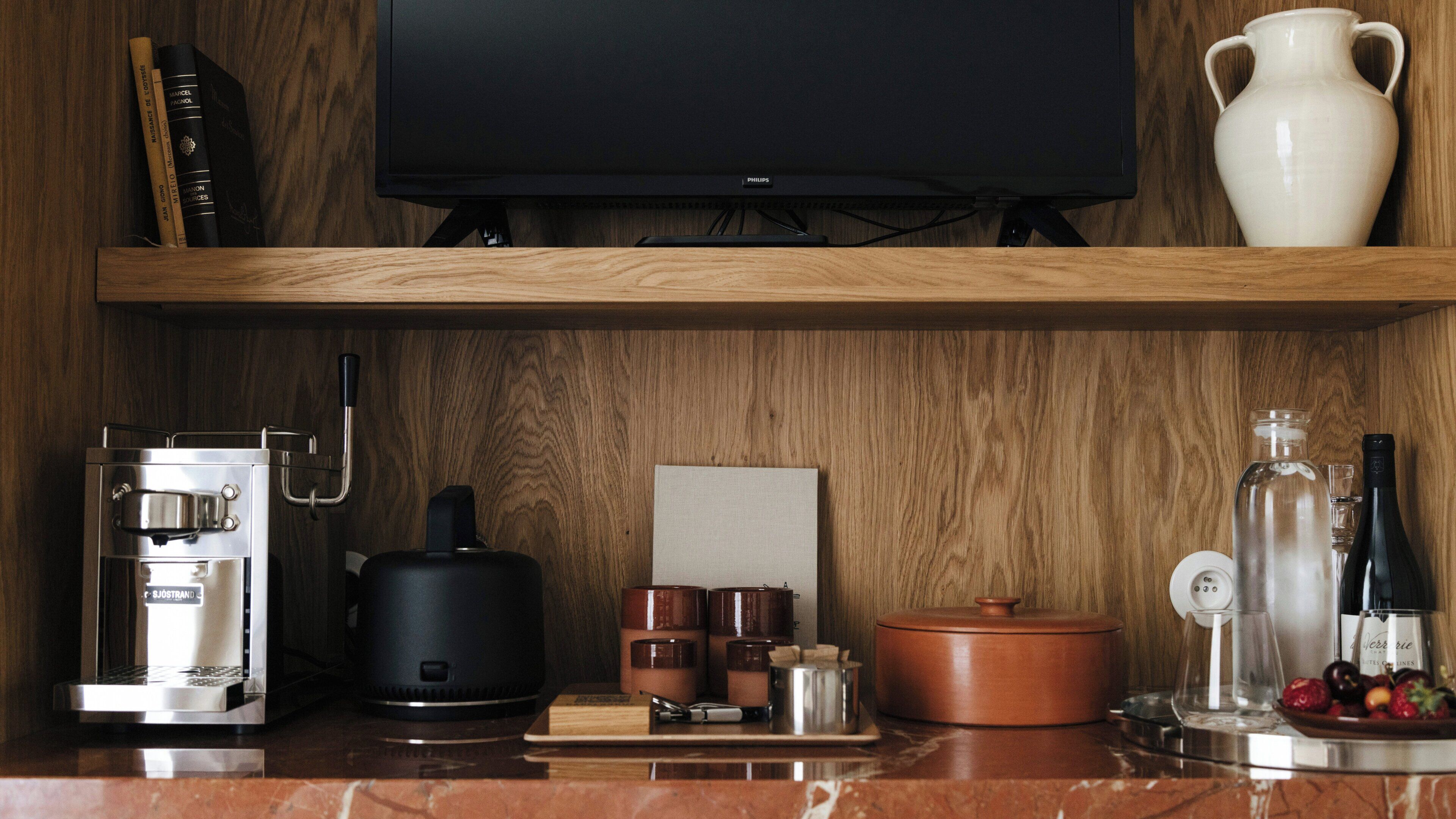 Una cocina con cafetera, televisor, un florero blanco y estante de madera.