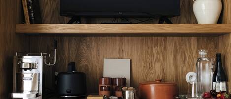 Una cocina con cafetera, televisor, un florero blanco y estante de madera.