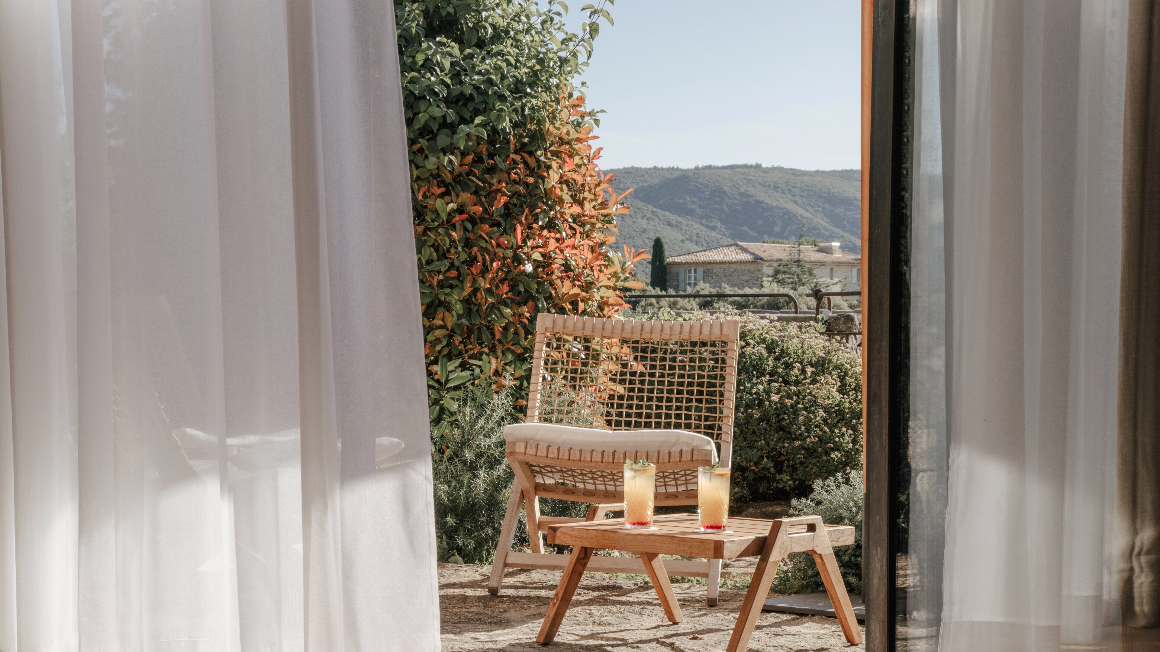 A wicker chair with a cushion and a glass of drink on a table, a view of a hill and buildings through a window with white curtains.