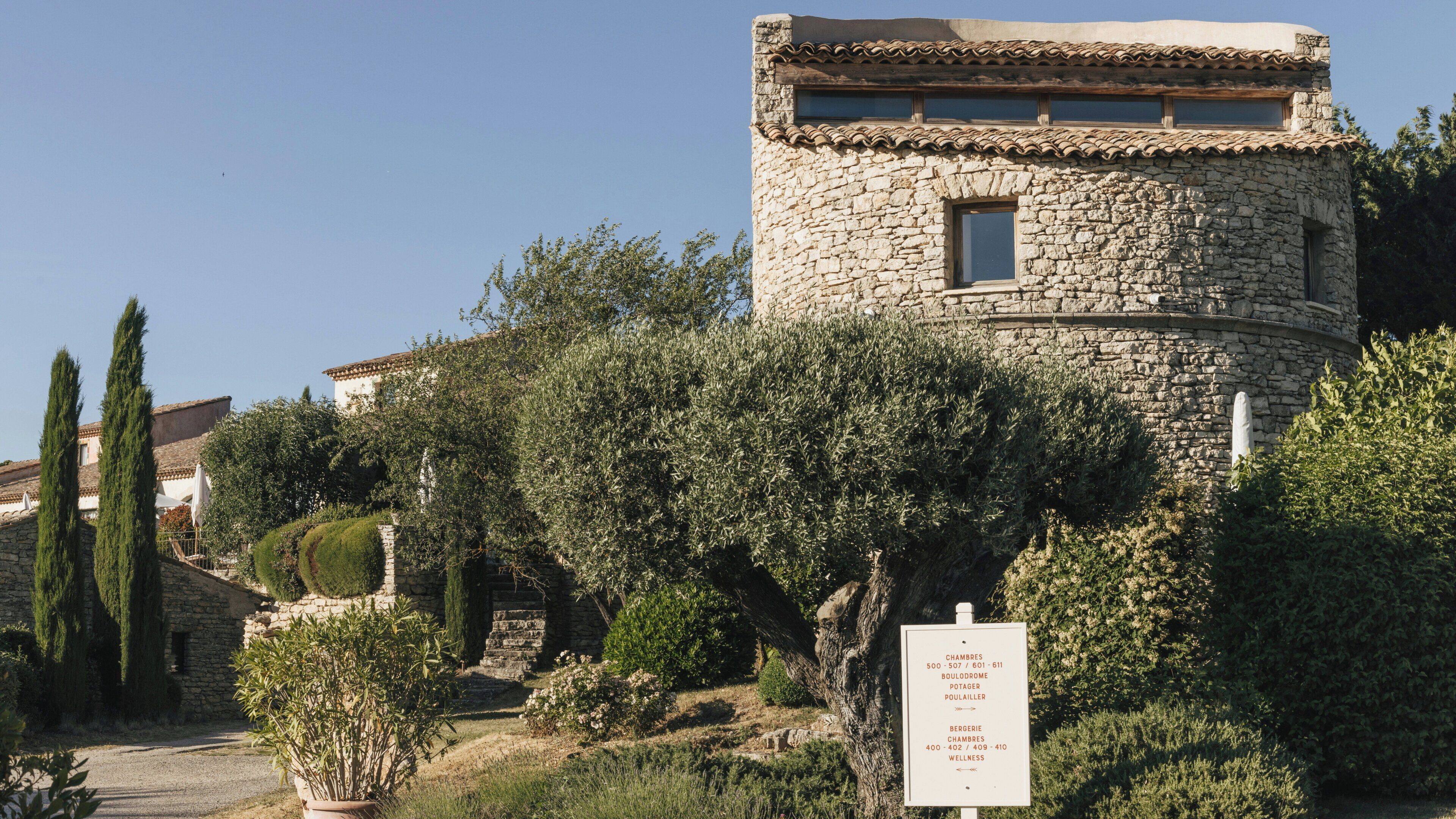 A stone building with a small window, surrounded by greenery and a sign listing room numbers and names.