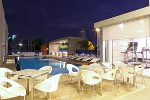 A swimming pool with lounge chairs and tables, surrounded by a modern building with a glass facade. - One Villahermosa 2000 (Villahermosa)