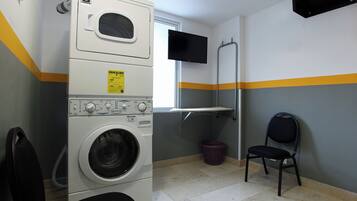 Une buanderie avec des machines à laver et des sèche-linge superposés, un téléviseur fixé au mur, une chaise et une petite fenêtre.