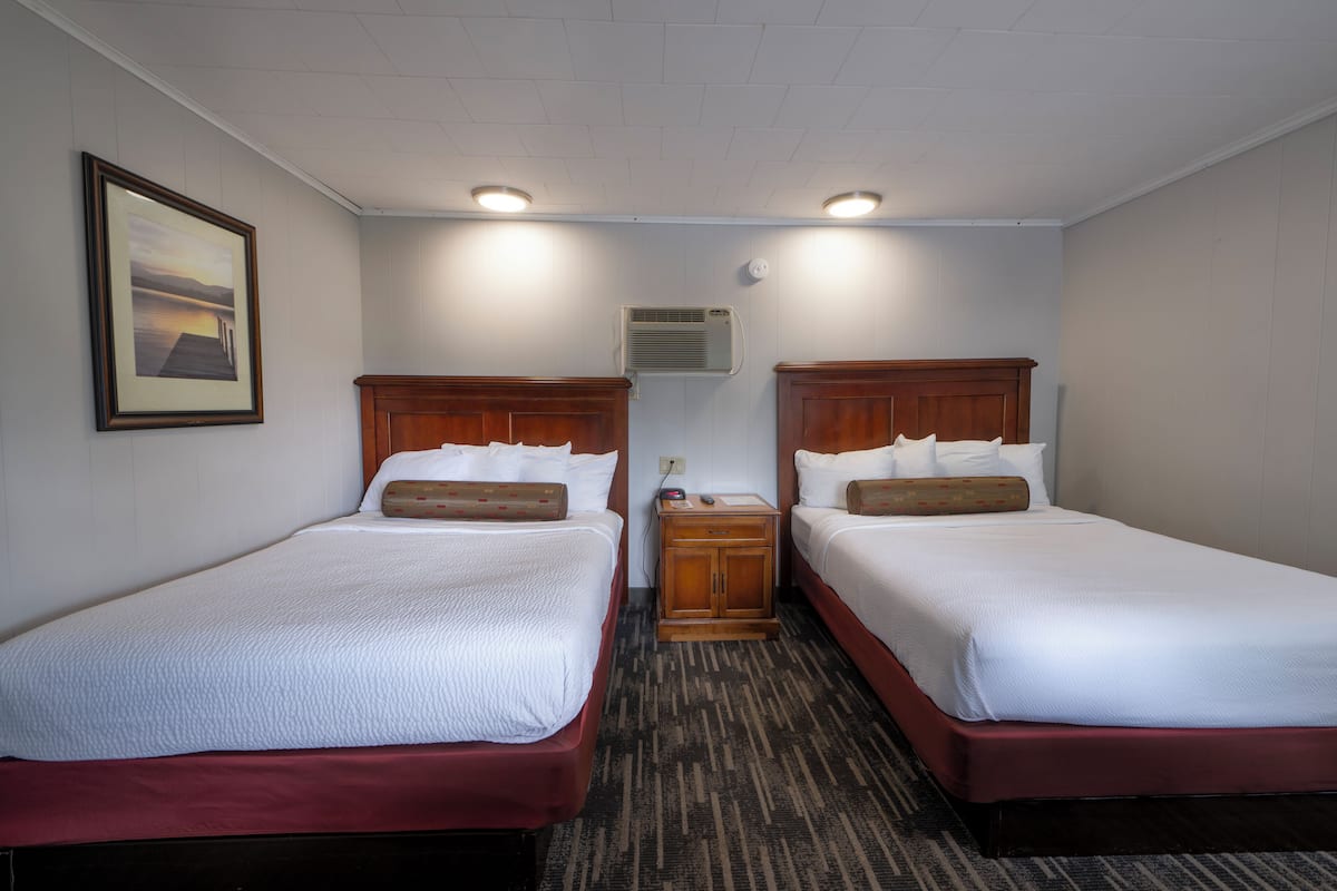 A hotel room with two beds, a wooden headboard, a framed picture on the wall, and an air conditioning unit.