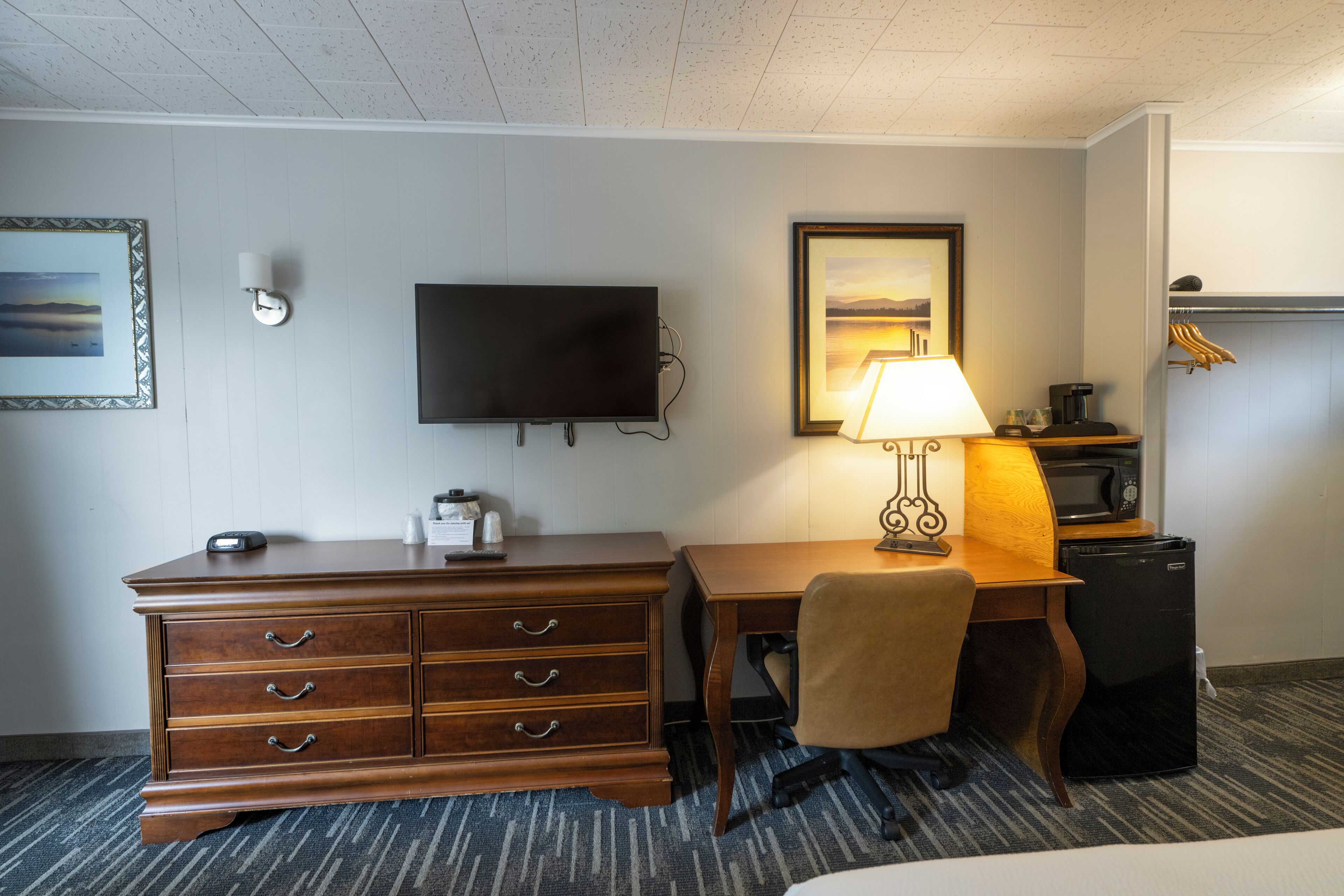 A hotel room with a wooden dresser, a desk with a lamp, a television, a microwave, and a small refrigerator.