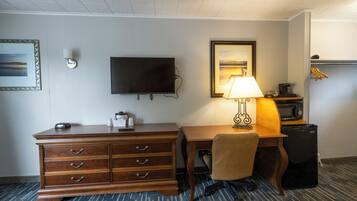 A hotel room with a wooden dresser, a desk with a lamp, a television, a microwave, and a small refrigerator.